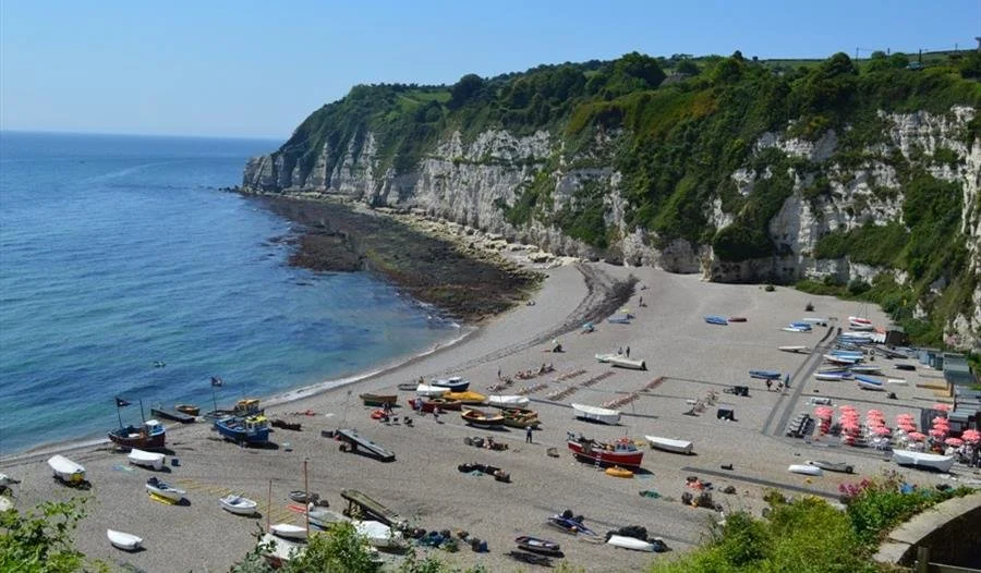 A scenic beach with boats, cliffs, and a small cove with umbrellas and lounge chairs.