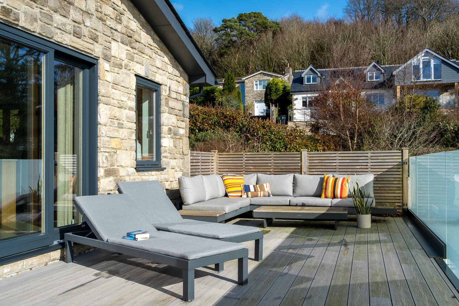 Outdoor patio area with lounge chairs, a sectional sofa with colorful striped pillows, a potted plant, and a wooden privacy fence. The house has stone exterior walls and sliding glass doors, with a backdrop of trees and neighboring houses.