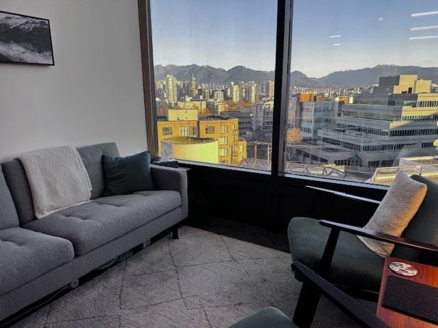 View of a city skyline with mountains in the background from an office window, with a gray sofa and an armchair in the foreground.