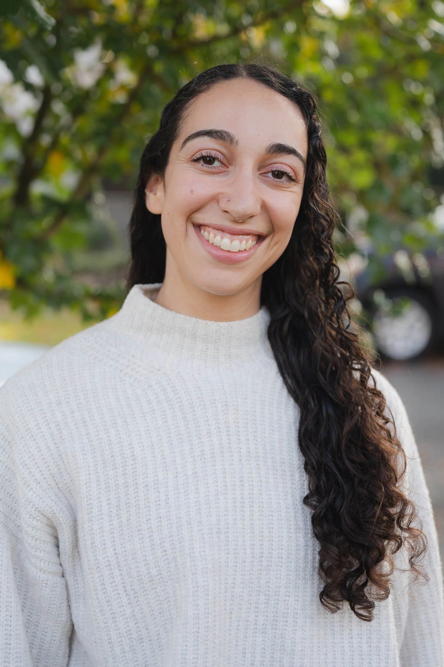 Close-up of a smiling woman with long curly dark hair, wearing a white knit sweater, outdoors with green trees in the background.