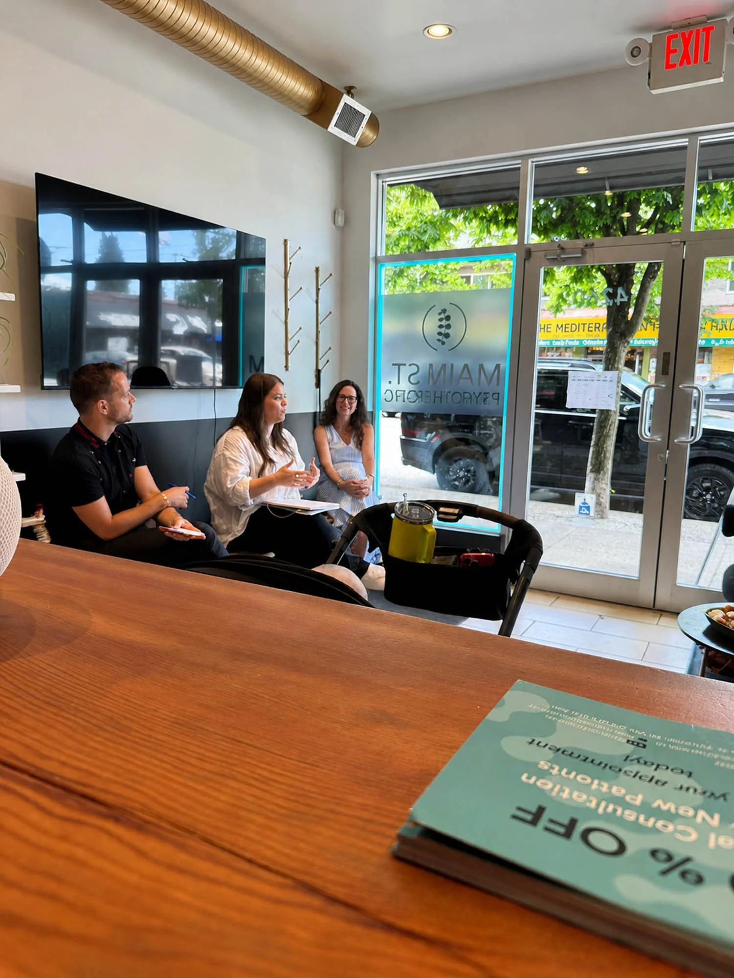 Three people sitting on a black bench in a modern cafe, talking and taking notes. A large television screen is mounted on the white wall behind them. The cafe has large glass doors with the cafe's name and logo, showing a street view with cars and a tree outside.