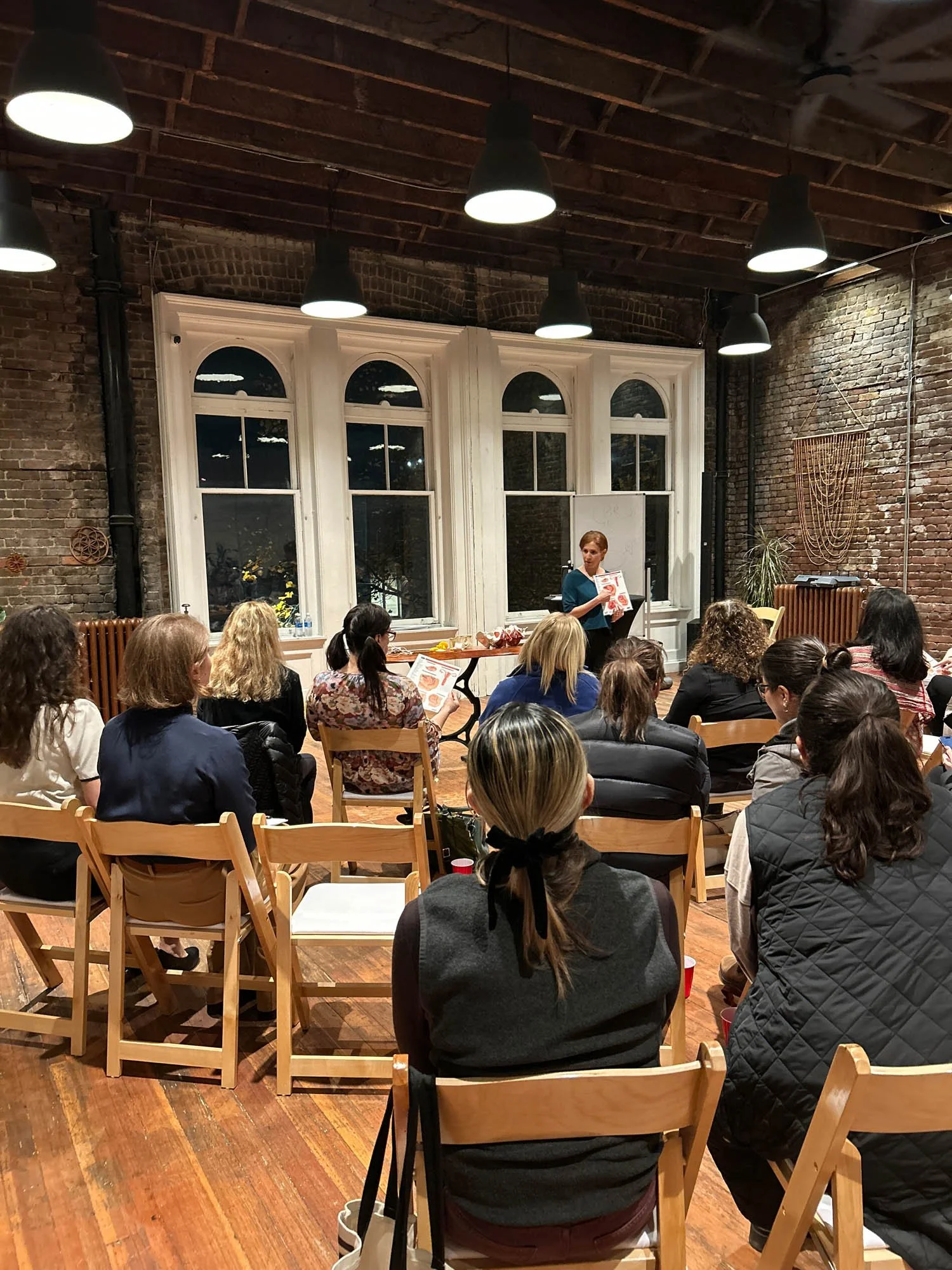 A woman speaking or presenting to an audience in a room with exposed brick walls, large windows, and wooden floors, with some audience members holding papers or pamphlets.