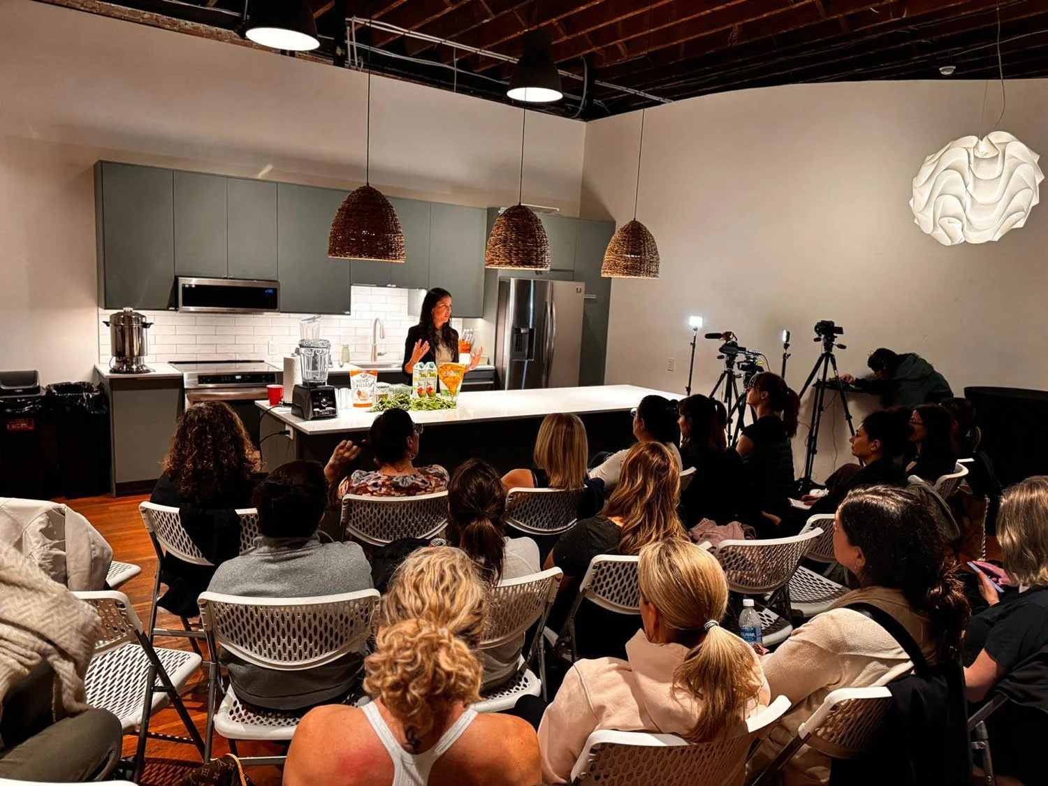 A woman presenter speaking to a seated audience in a modern kitchen studio, with cameras and lighting equipment set up for recording.