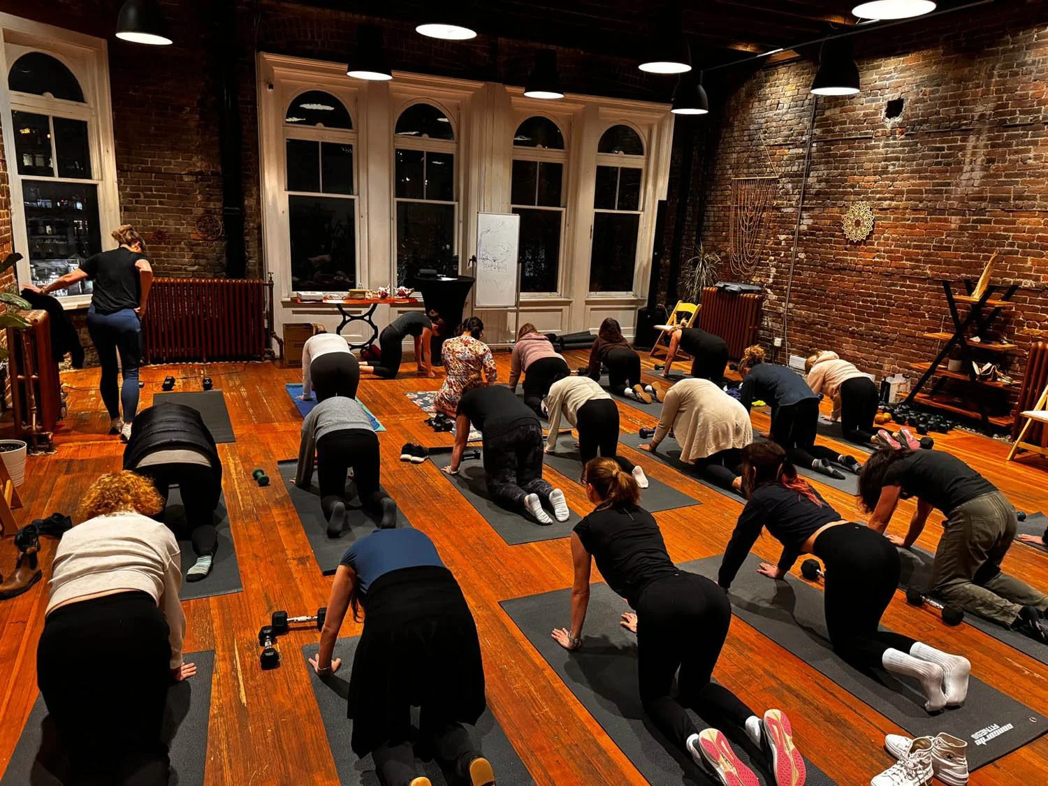 People participating in a yoga class in a spacious room with brick walls and large windows, practicing a downward dog pose.