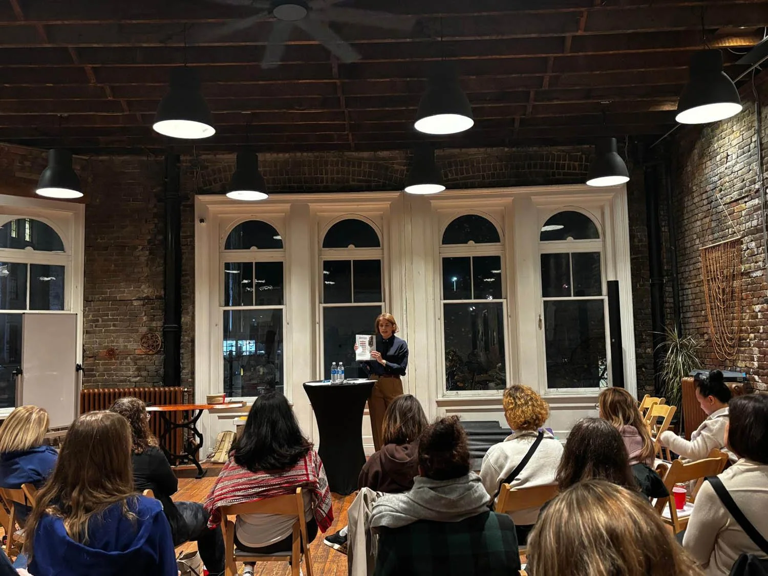 A woman is giving a presentation in a room filled with an audience of women. The room has brick walls, large arched windows, and is lit by ceiling lights. The speaker stands at a tall table holding a paper, with water bottles on the table.