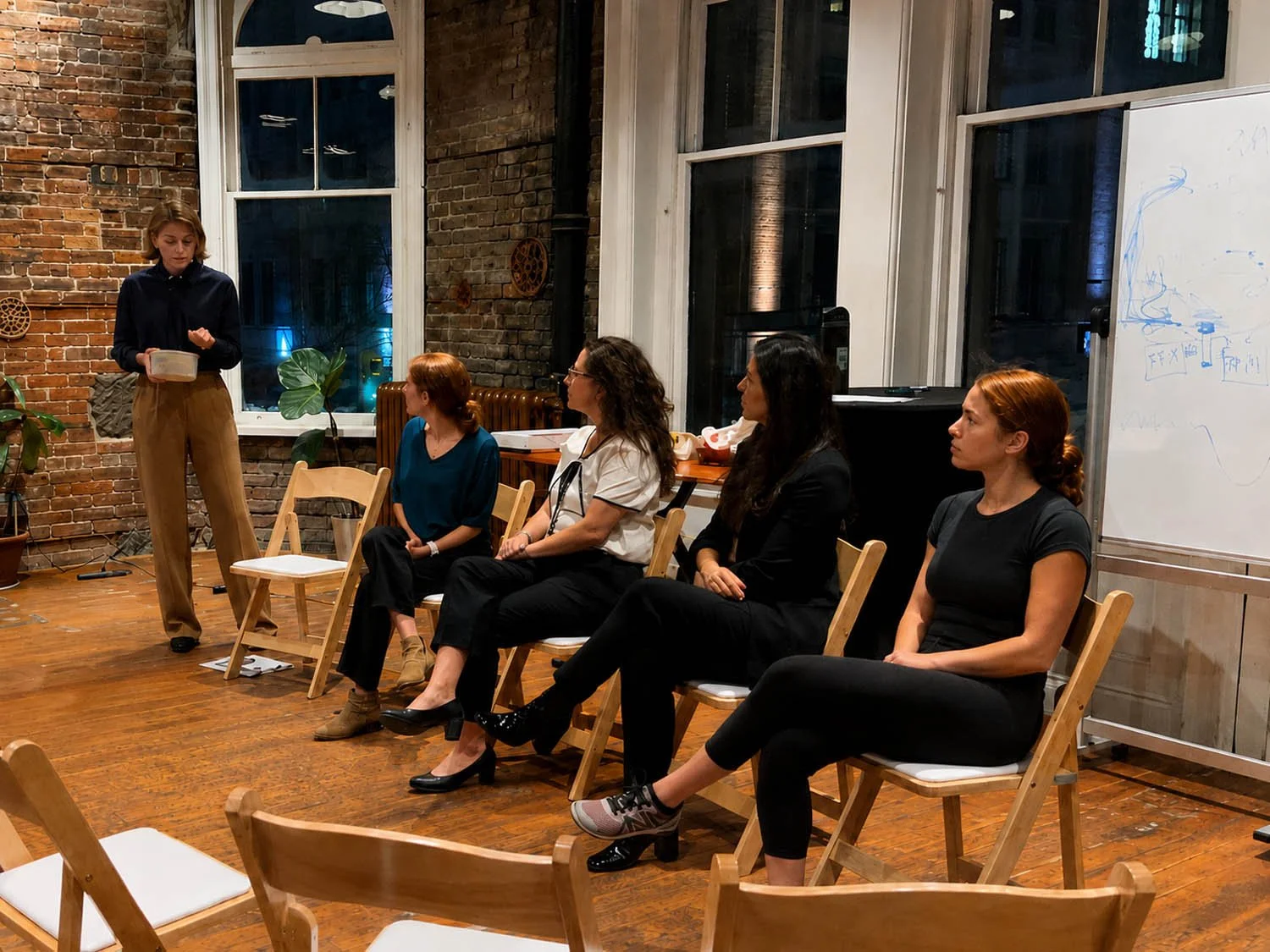 A woman is standing and speaking to four seated women in a room with brick and glass walls and wooden floors, possibly a workshop or seminar setting.