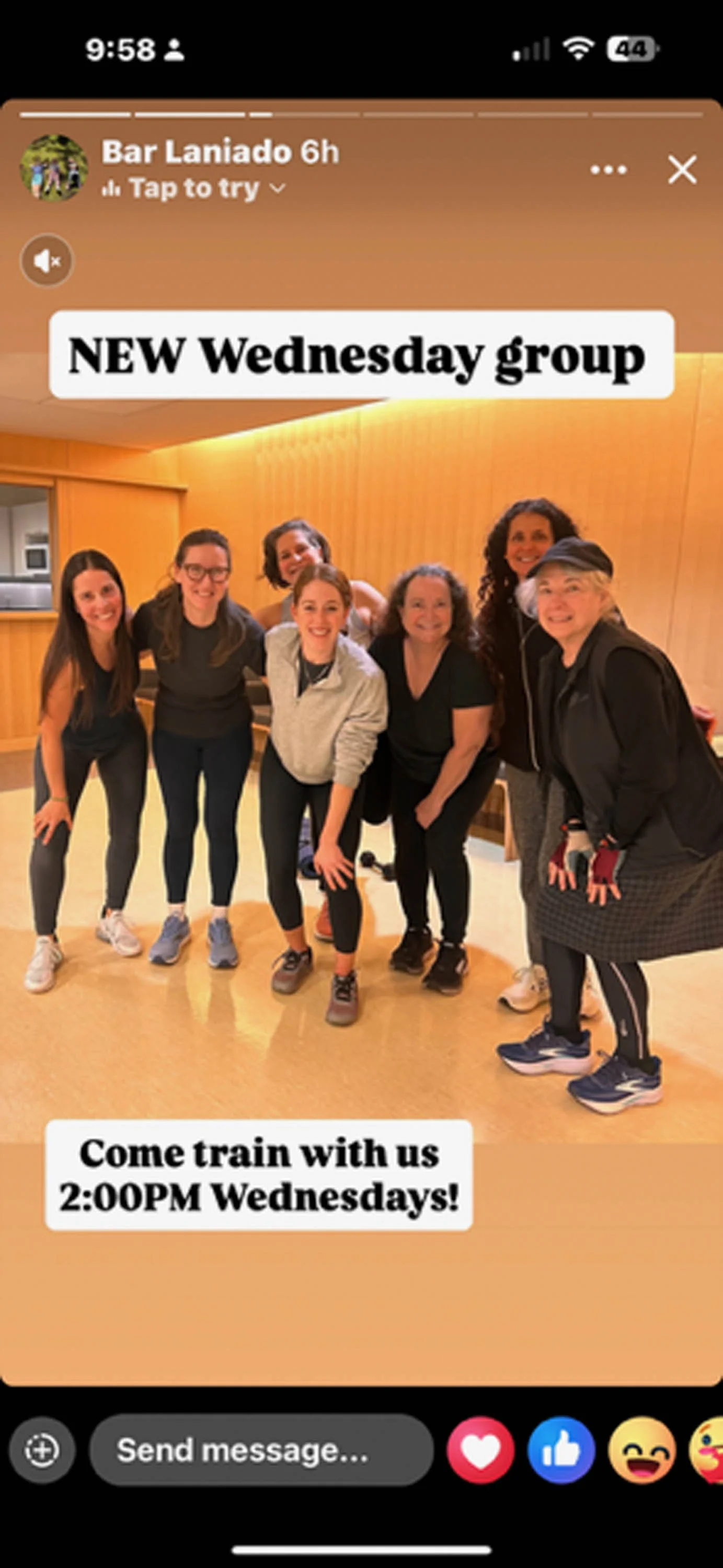 A group of seven women gathered in a gym or fitness studio, smiling and posing together for a photo. The background features warm lighting and wooden paneling.