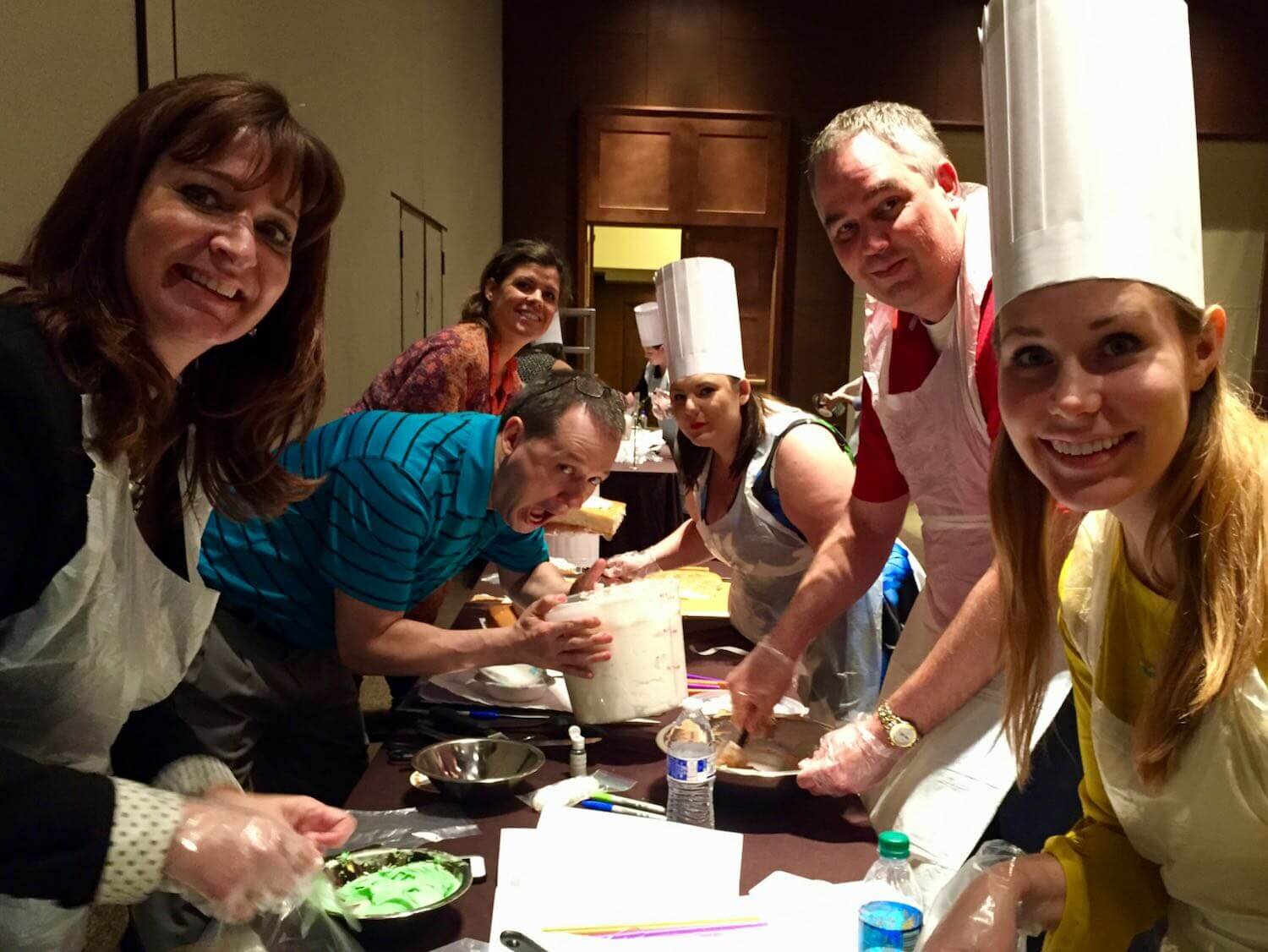Group of people cooking and smiling in a kitchen setting, wearing chefs' hats and aprons.