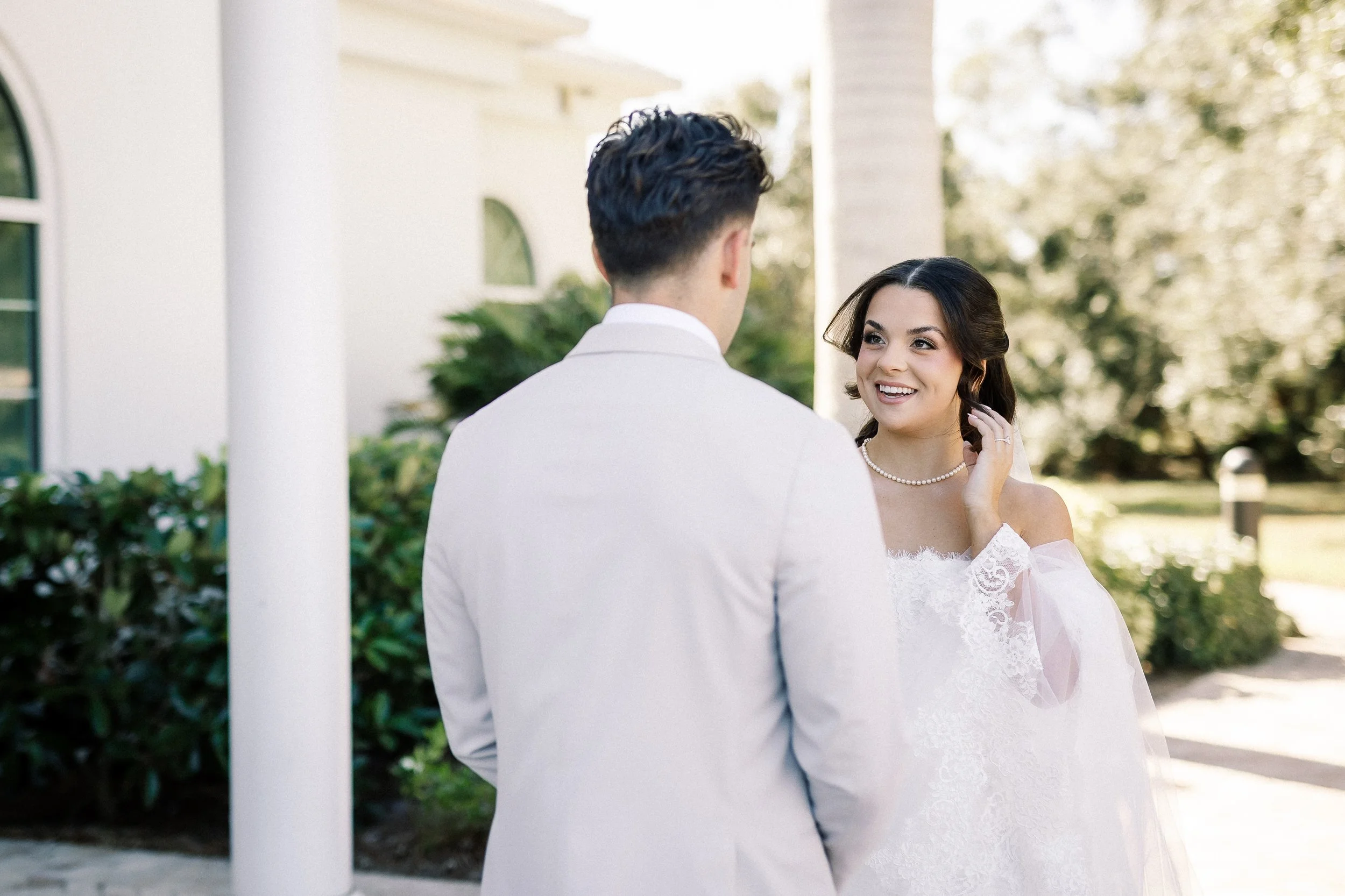A bride and groom during their wedding ceremony outside, with the bride smiling and looking at the groom, surrounded by greenery and a white building.
