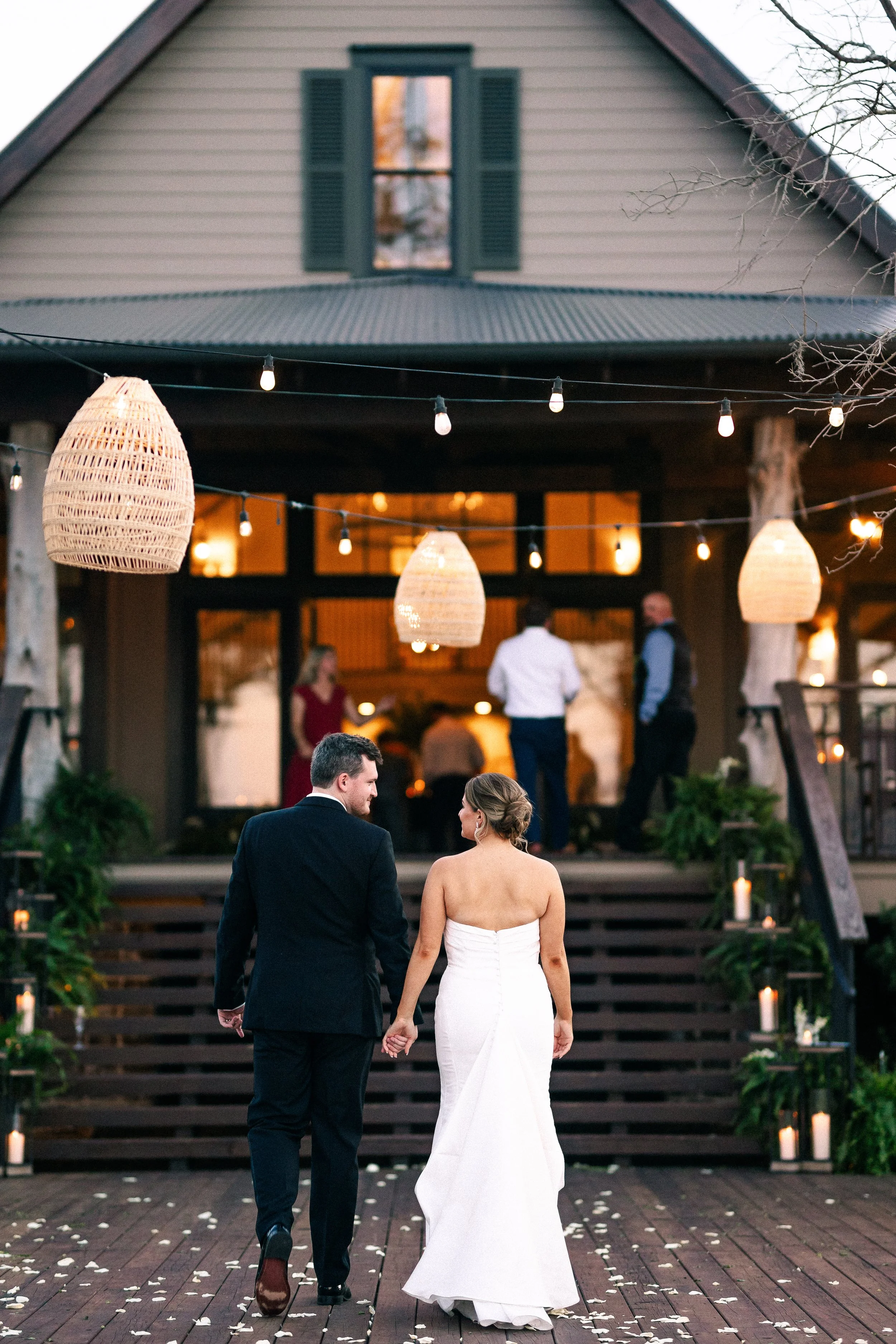 A bride and groom walking hand in hand on a wedding day, approaching a house decorated with hanging lights and candles on the stairs, with guests in the background.