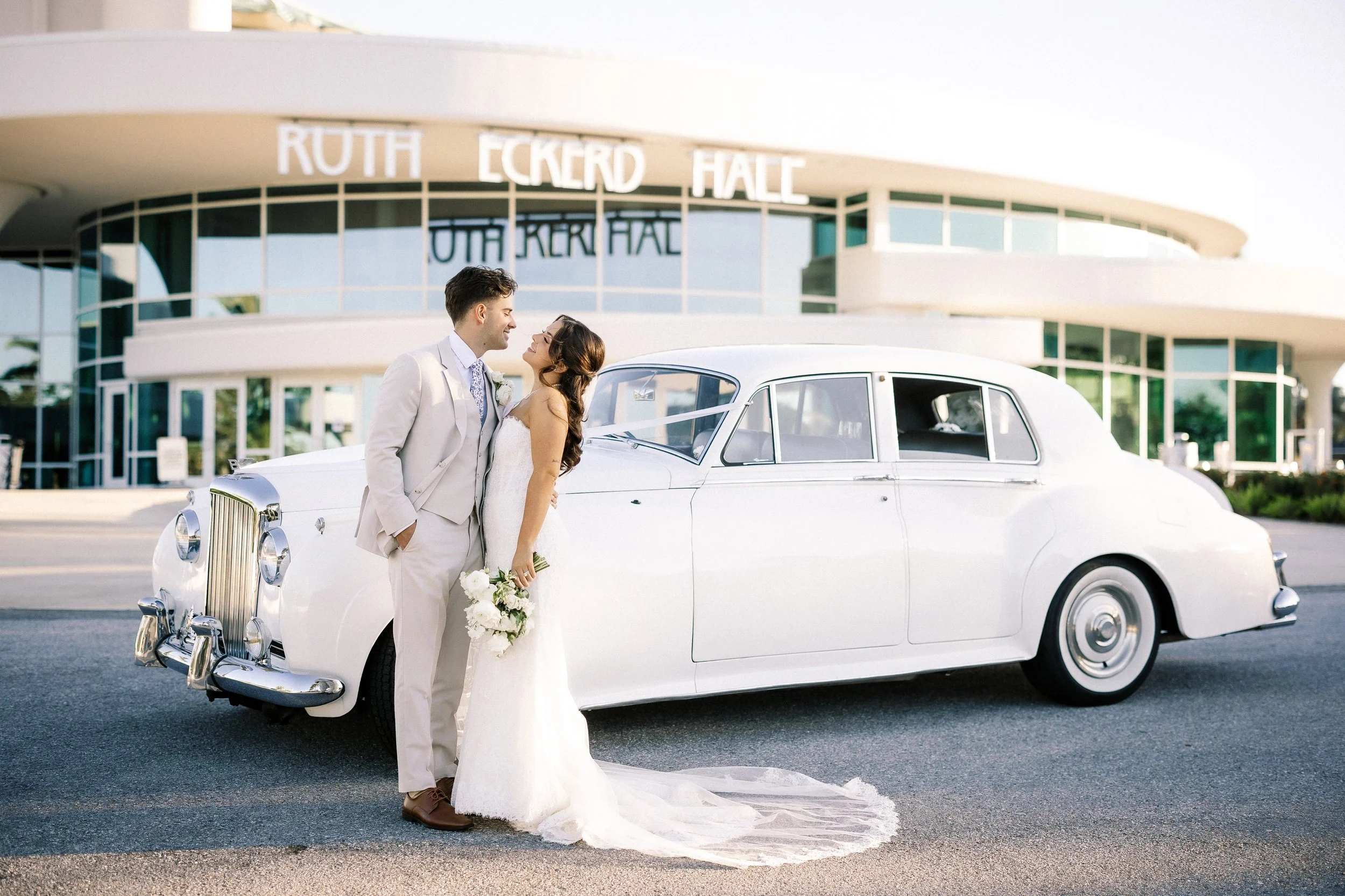 A bride and groom in wedding attire standing in front of a white vintage car outside a building with a sign for Ruth Eckerd Hall.