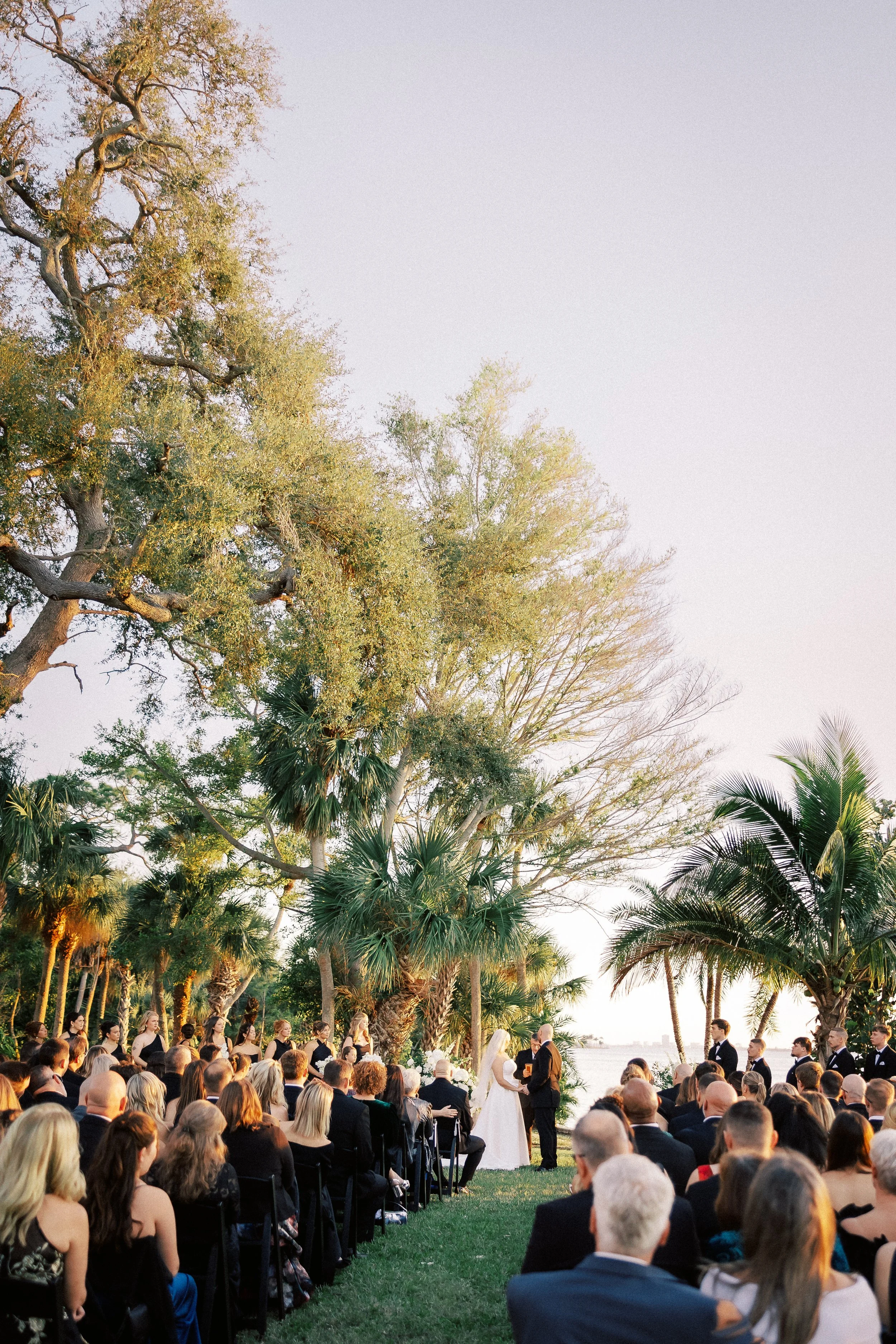 Outdoor wedding ceremony with guests seated on grass, bride and groom exchanging vows by a lake, surrounded by palm trees and large trees, during sunset.