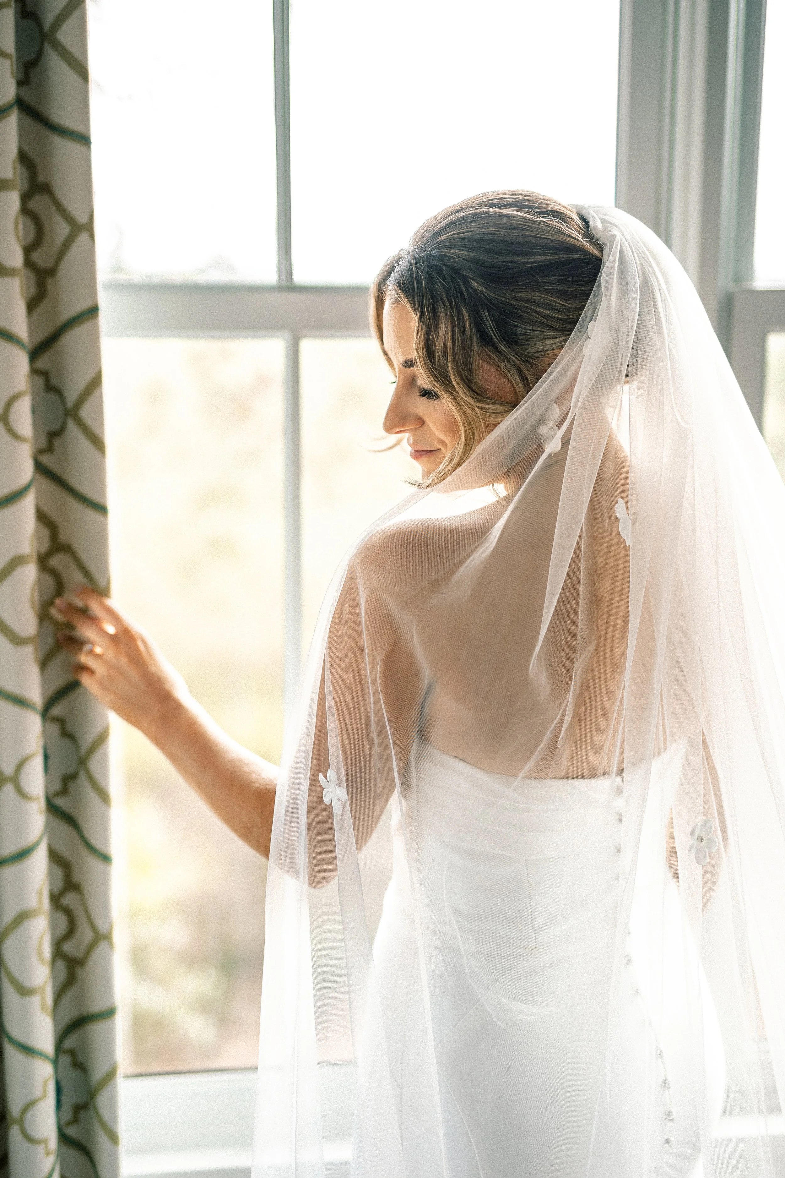 Bride standing by a window with sunlight streaming in, wearing a wedding dress and veil, looking down, with a thoughtful expression.