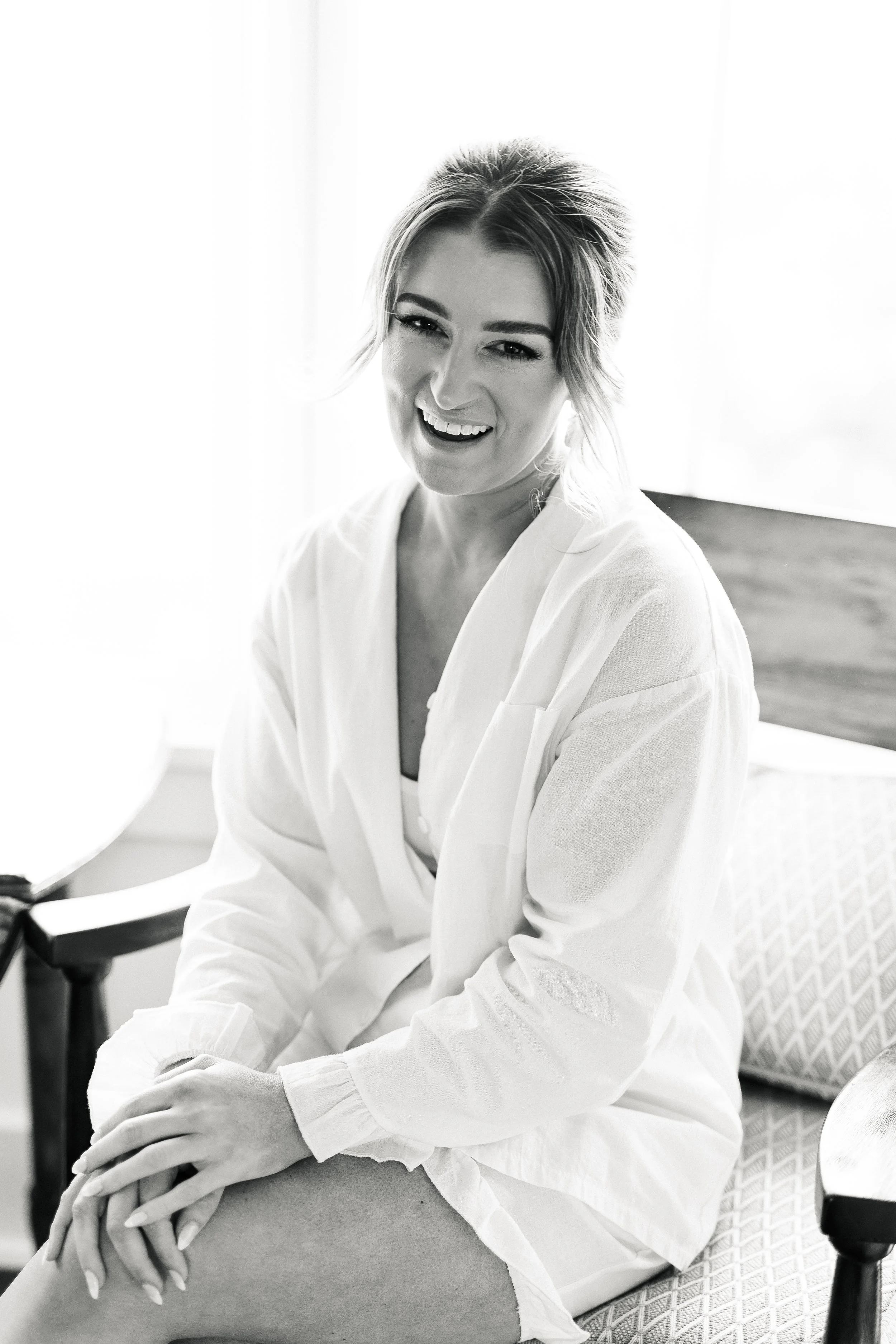 Black and white photo of a woman with short hair, smiling, sitting on a chair, wearing a light-colored blouse or dress.