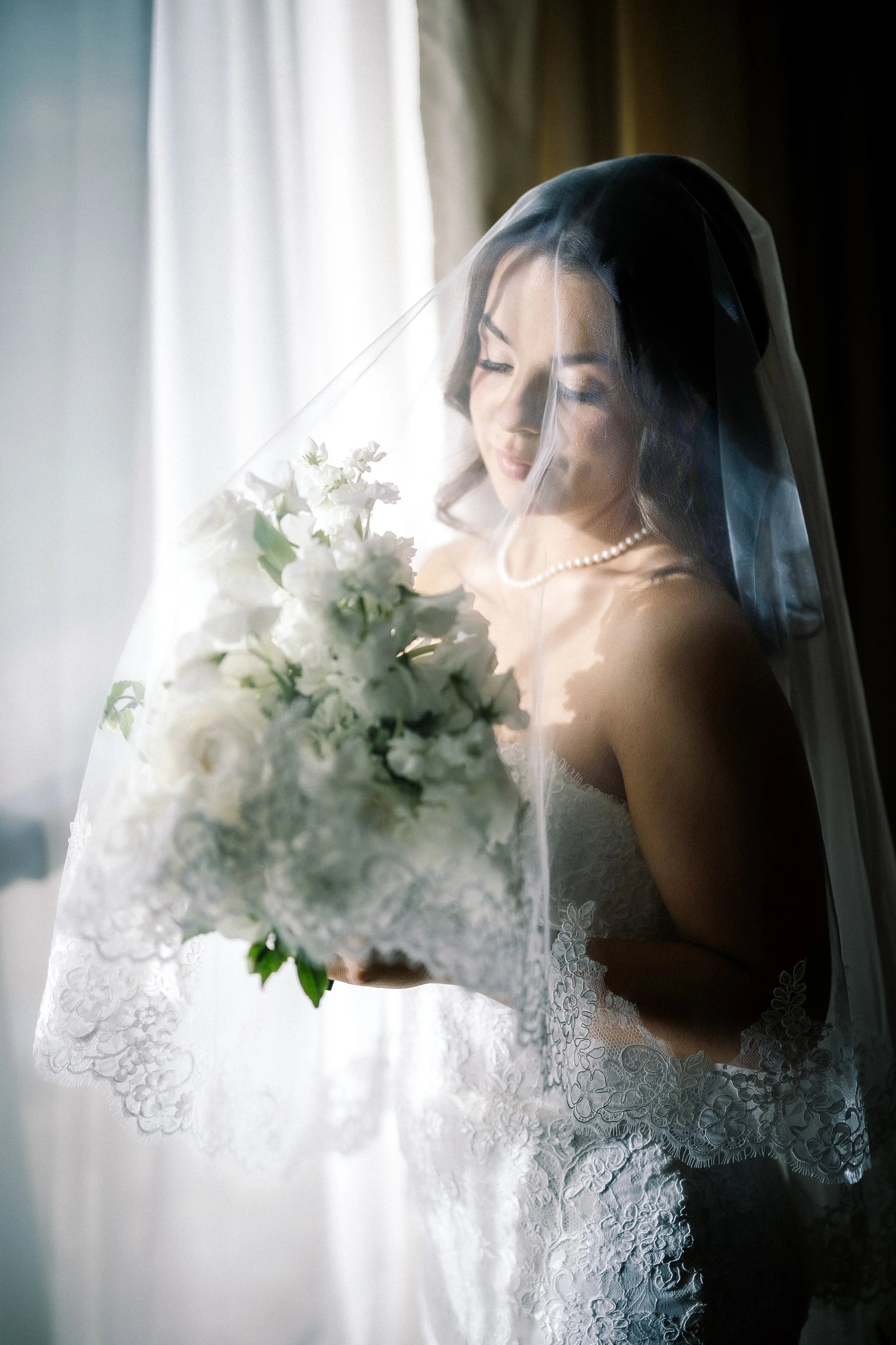 Bride in wedding dress holding bouquet, looking at flowers through sheer curtain, soft natural light.