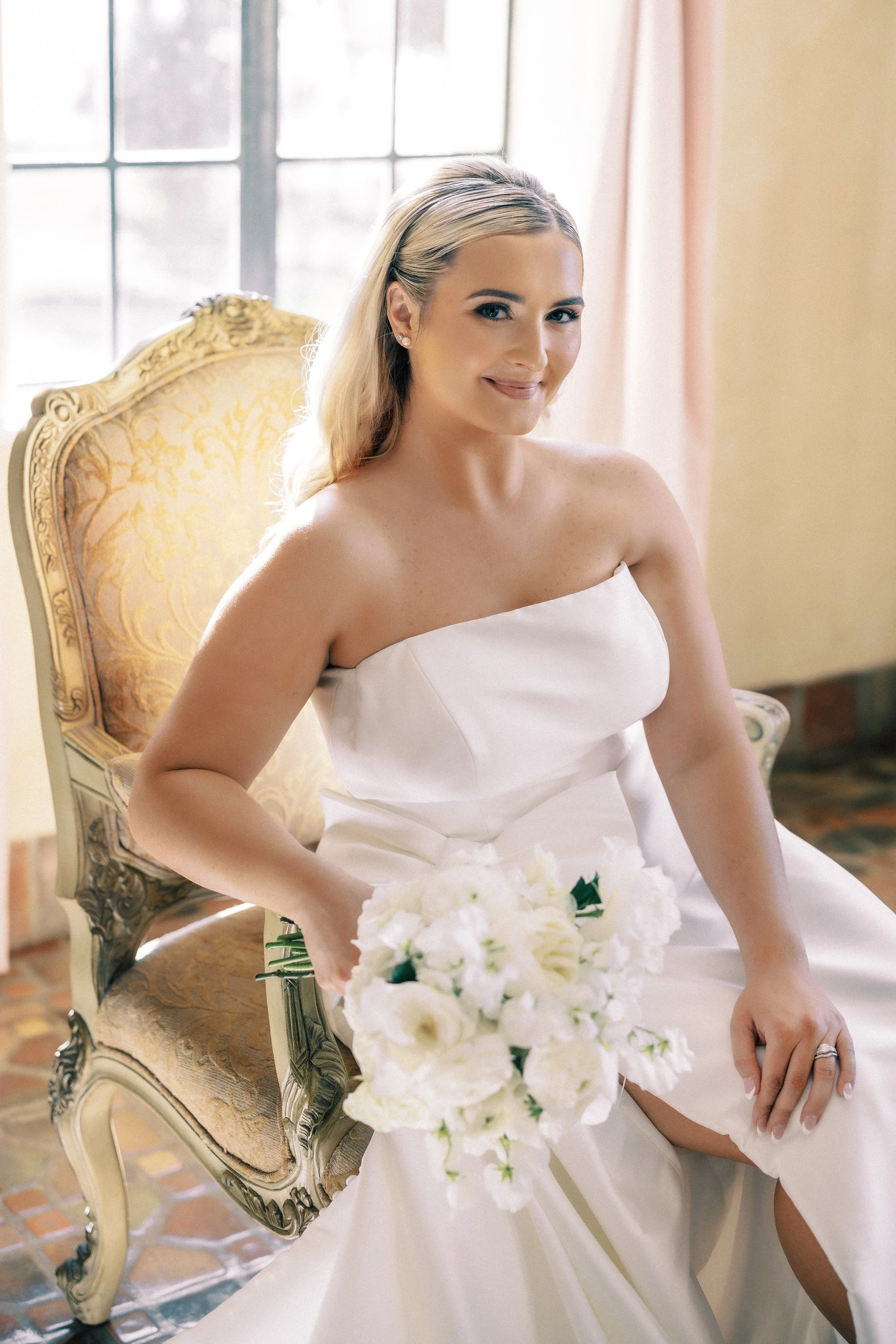 A bride sitting on an ornate, vintage chair holding a bouquet of white flowers, wearing a strapless white wedding dress.