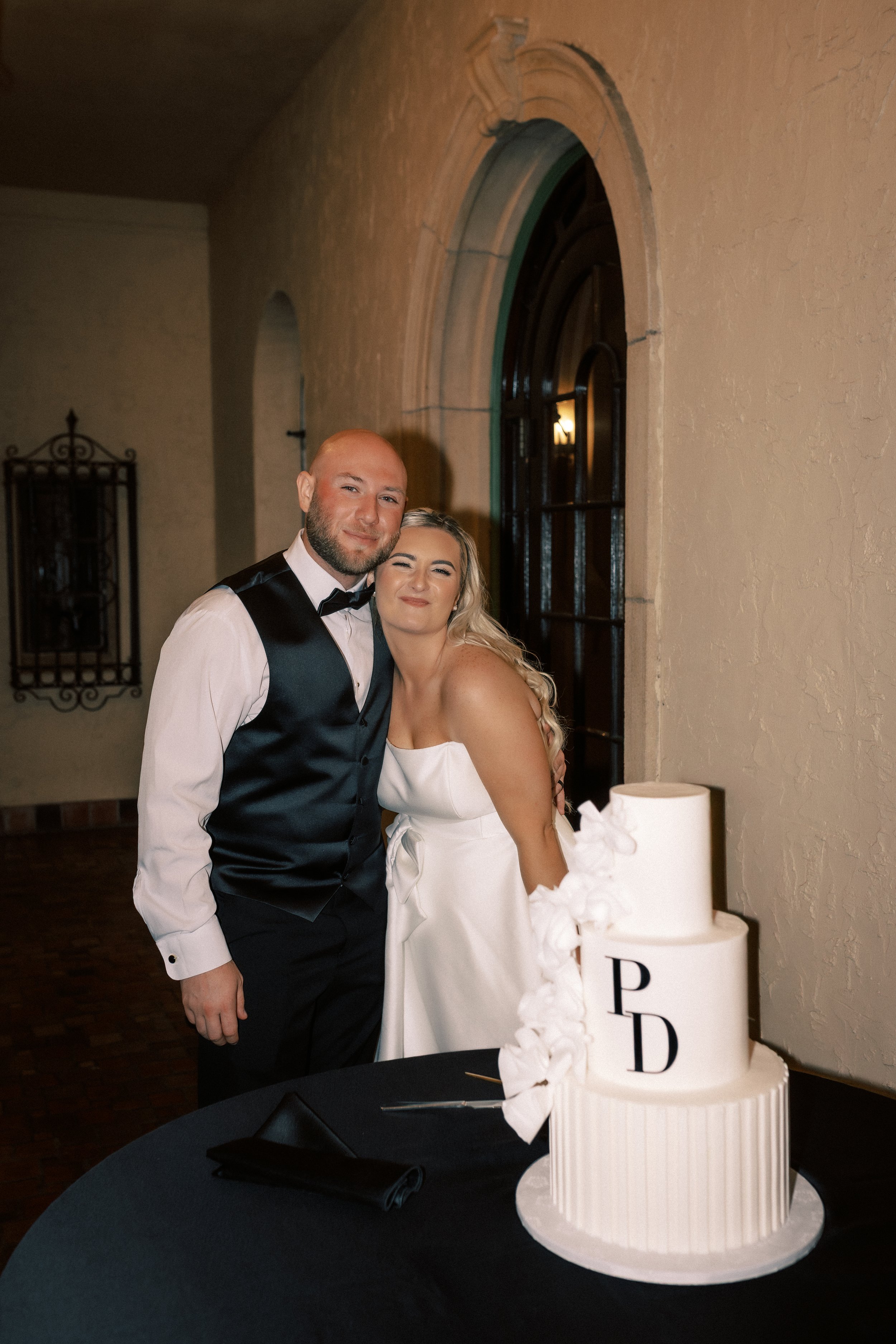 A newlywed couple, the groom in a black vest and bow tie, and the bride in a white strapless wedding dress, standing close together and smiling at a wedding reception. There is a three-tier white wedding cake with monogram initials 'P' and 'D' on the