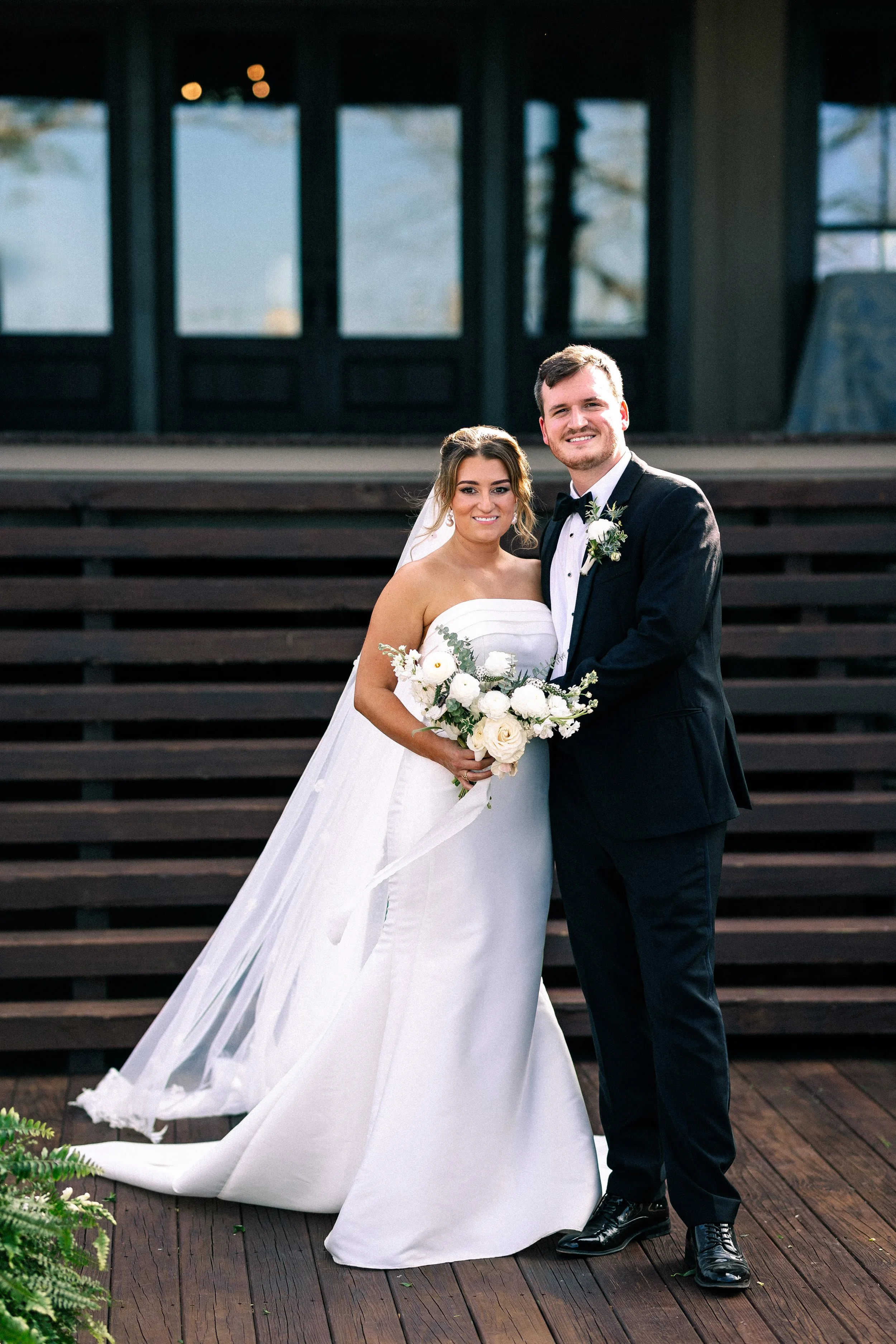 A bride and groom standing together outdoors, smiling. The bride is wearing a strapless white wedding gown with a long veil, holding a bouquet of white flowers. The groom is dressed in a black tuxedo with a white shirt, black bow tie, and a boutonnie