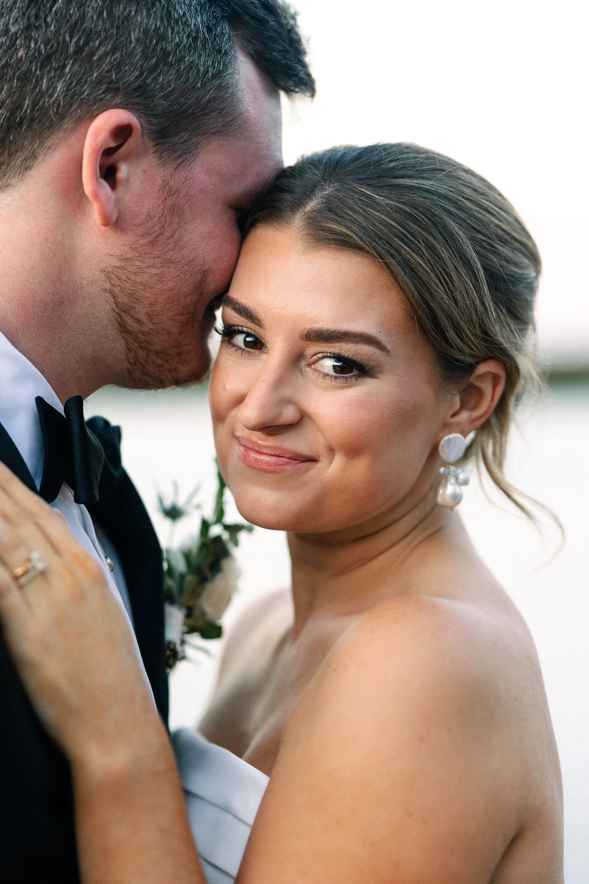 A newlywed couple close together, with the man whispering into the woman's ear and she smiling, outdoors during sunset.