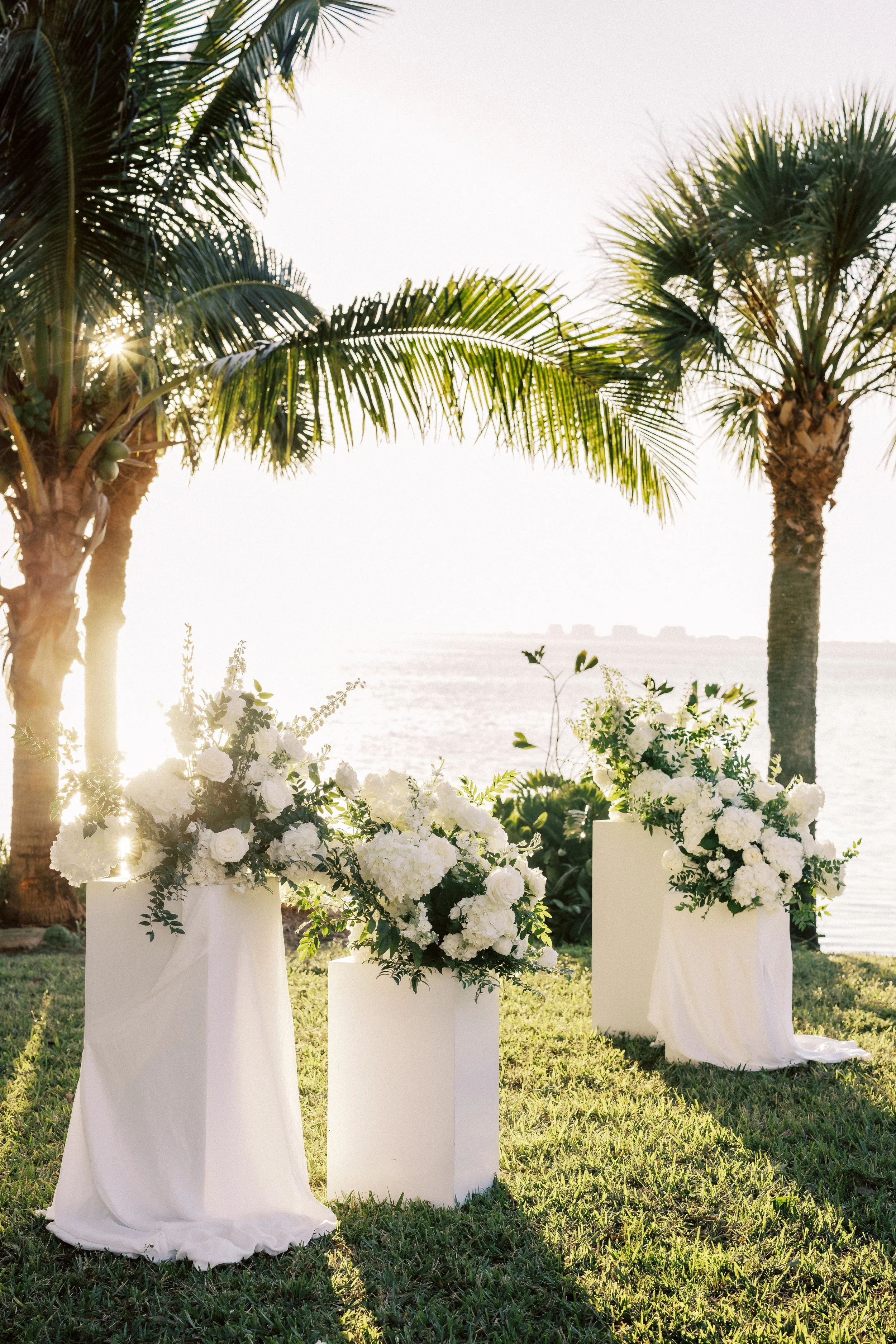 Three white pedestals with white floral arrangements on a grassy lawn by the water, palm trees in the background, sunlight shining through leaves at sunset.