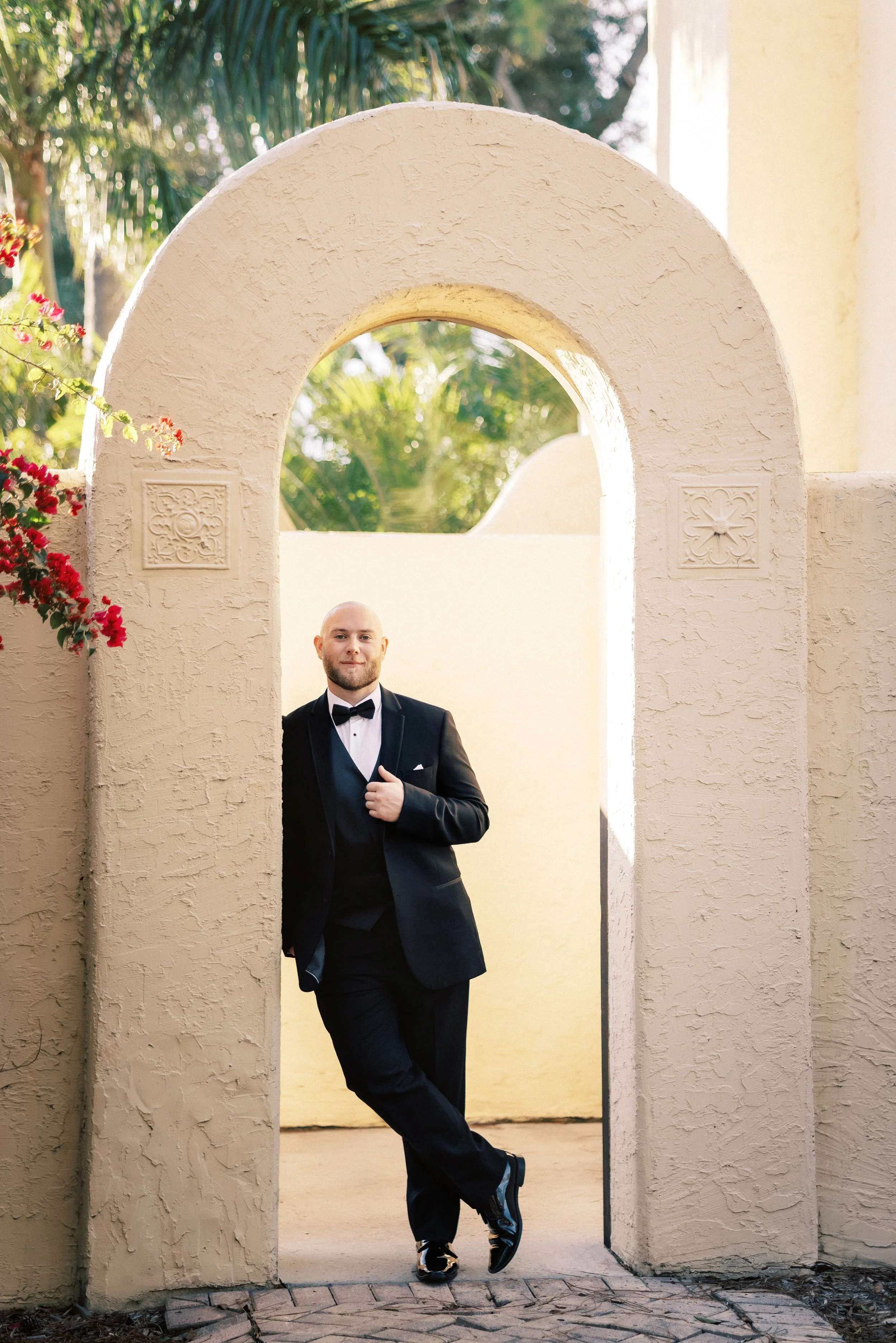 A man in a tuxedo standing under an archway with greenery in the background.