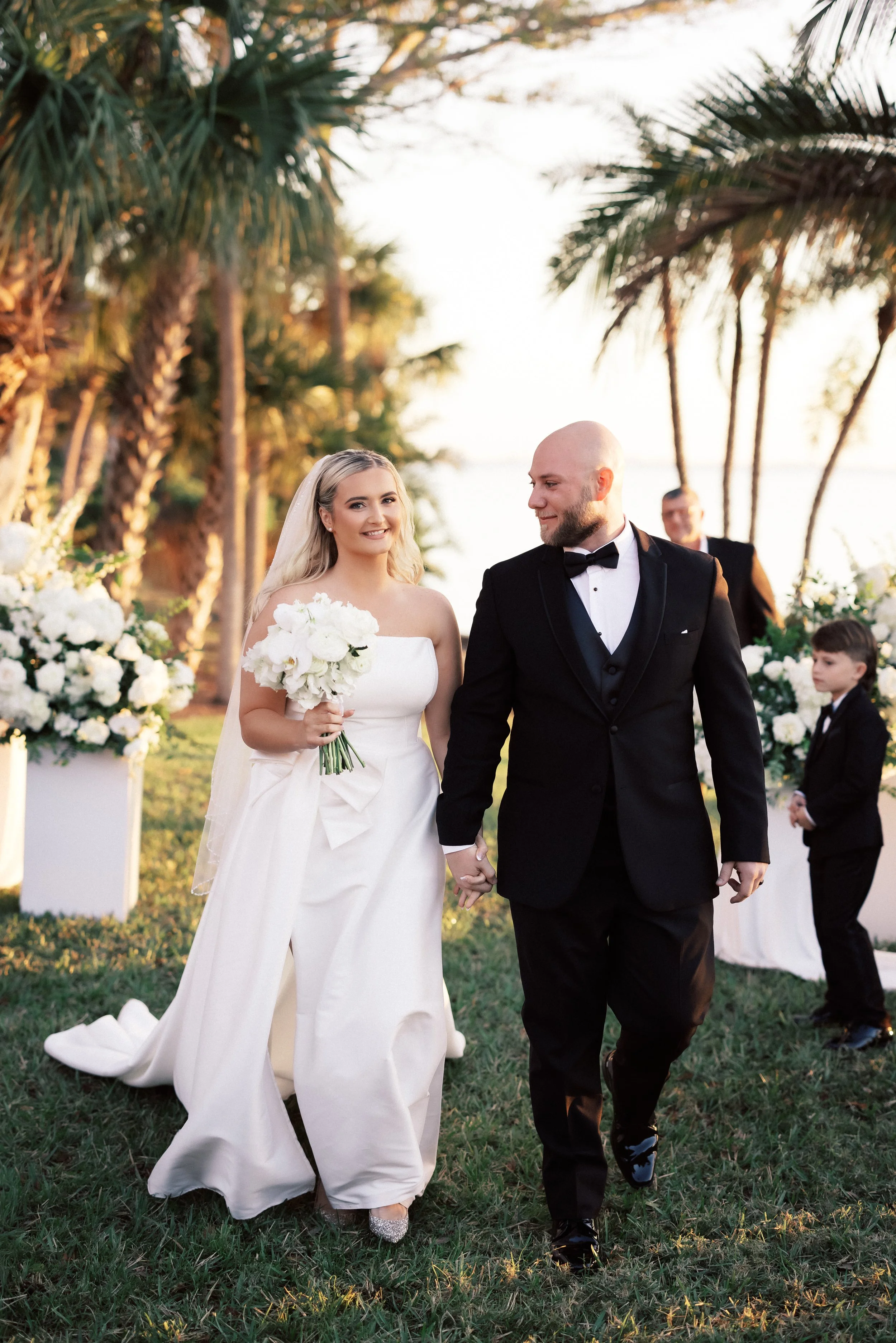 A bride and groom walking hand in hand outdoors during their wedding, surrounded by large white floral arrangements and tall palm trees, with a sunset in the background.