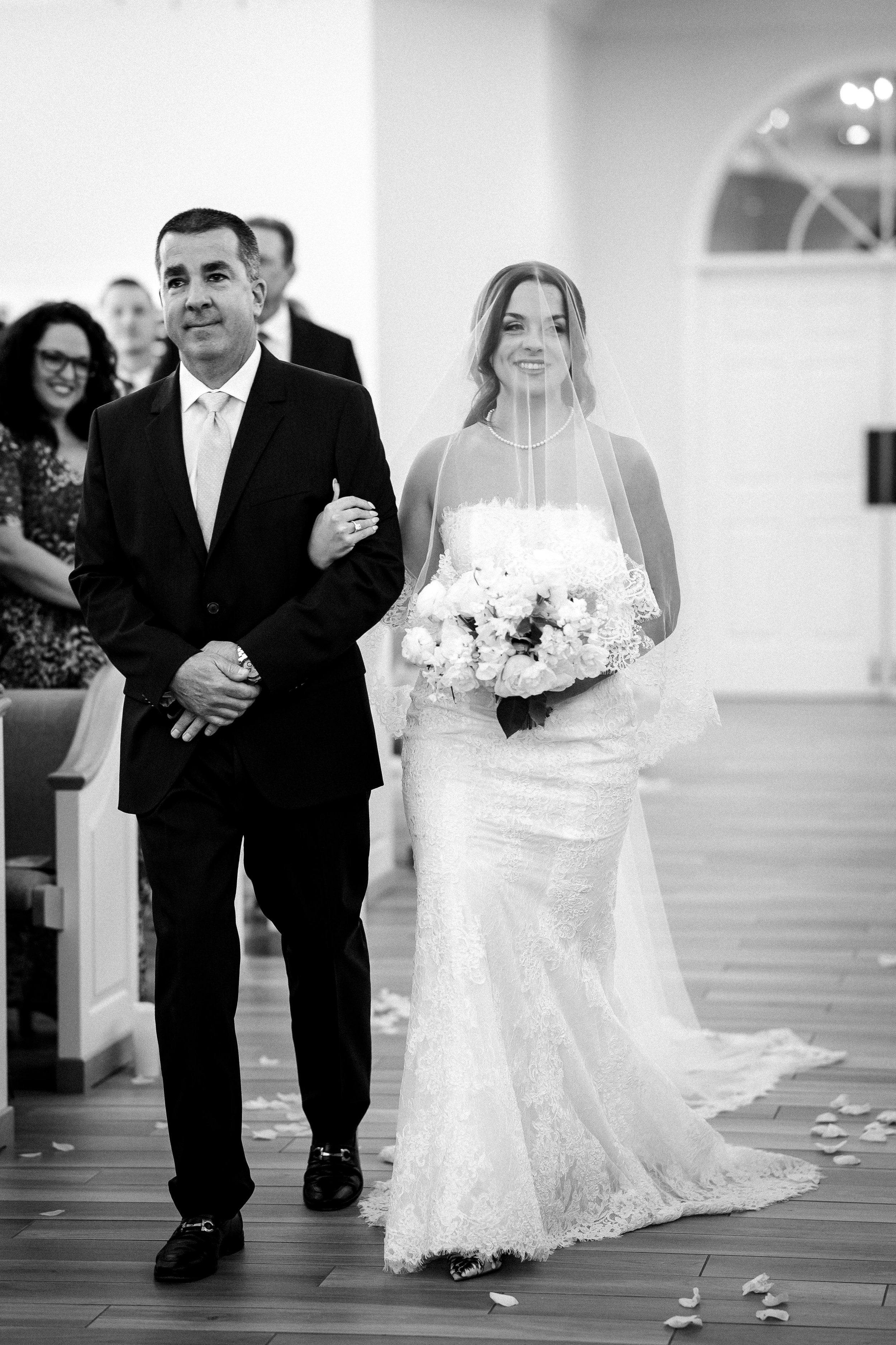A bride walking down the aisle with her father in a wedding ceremony. The bride is holding a bouquet of flowers, wearing a lace wedding dress and veil, and smiling. The father is dressed in a dark suit with a tie, and there are guests seated and smil