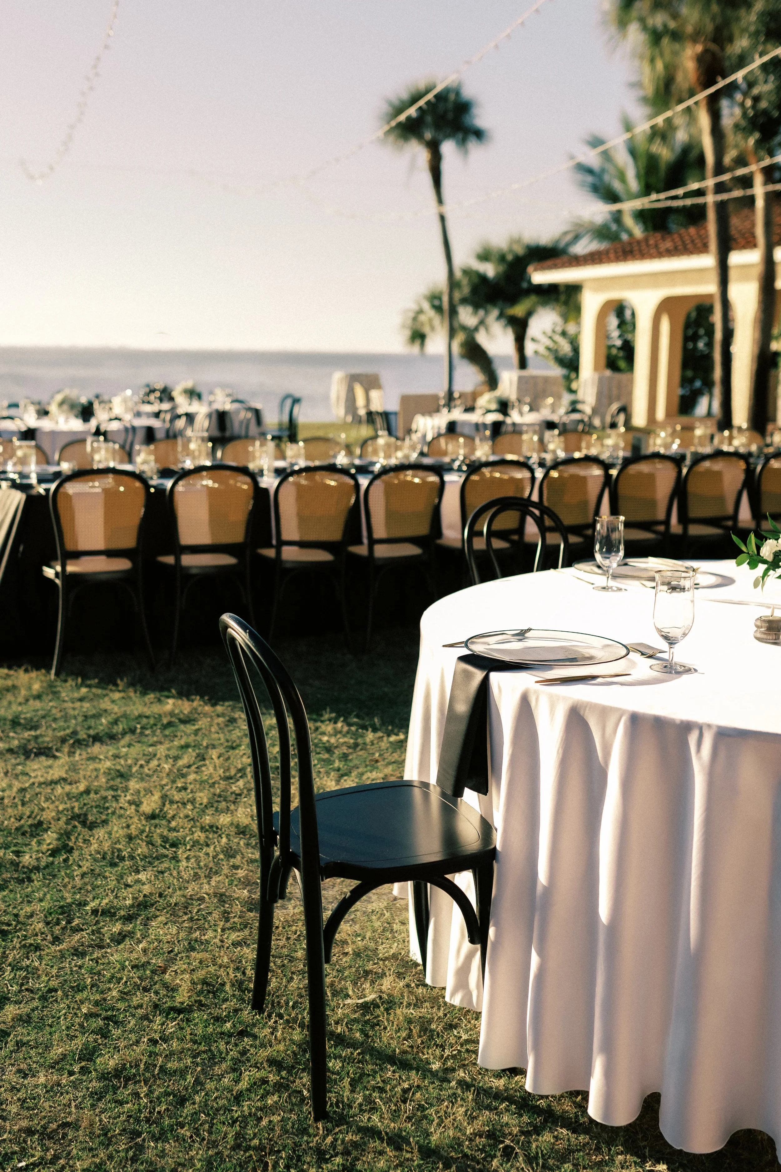 Outdoor event setup with round tables covered in white tablecloths, set with glassware and plates, under string lights near palm trees by the water at sunset.