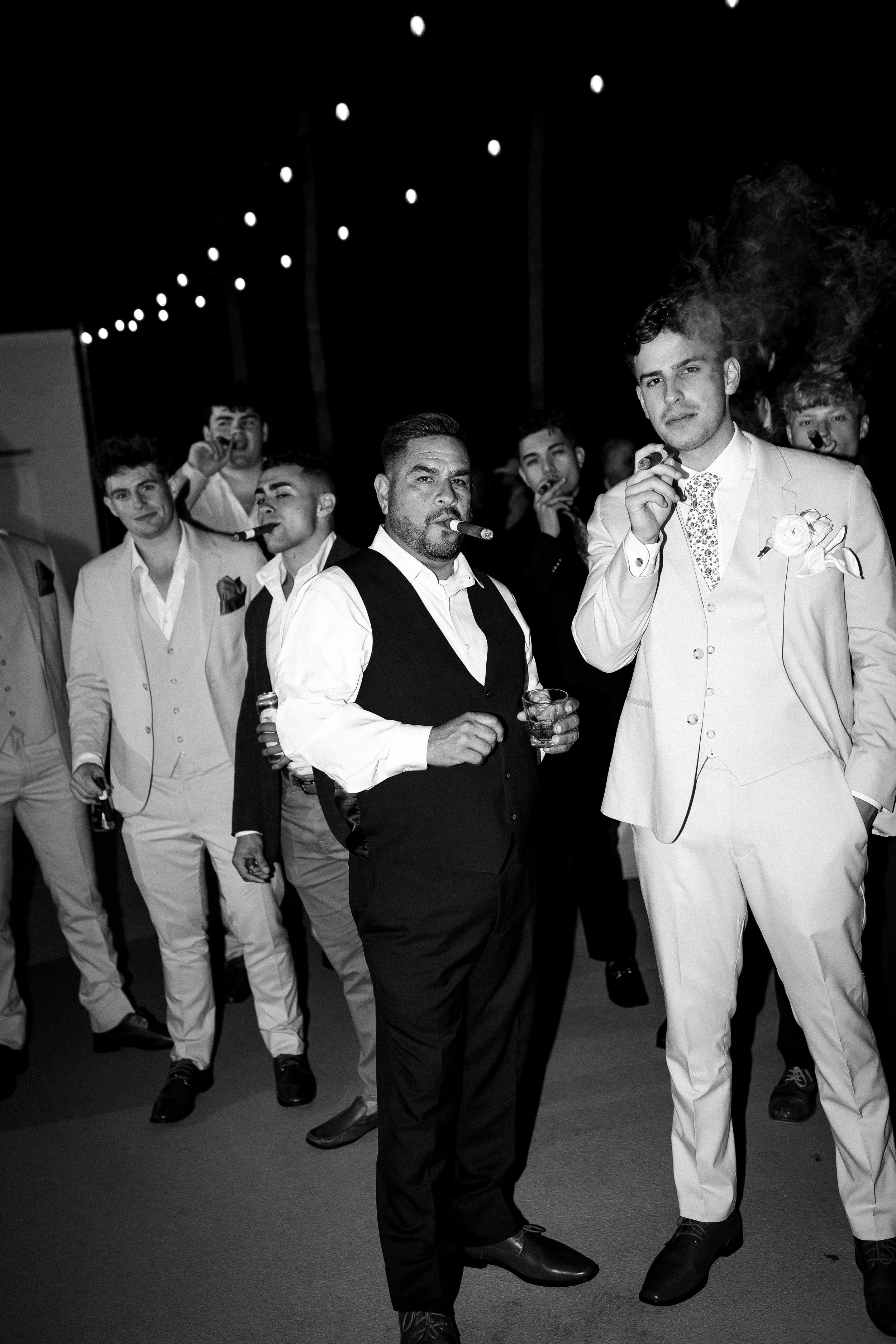 Group of men dressed in formal suits smoking cigars at a celebration or party, with string lights above and a dark background.