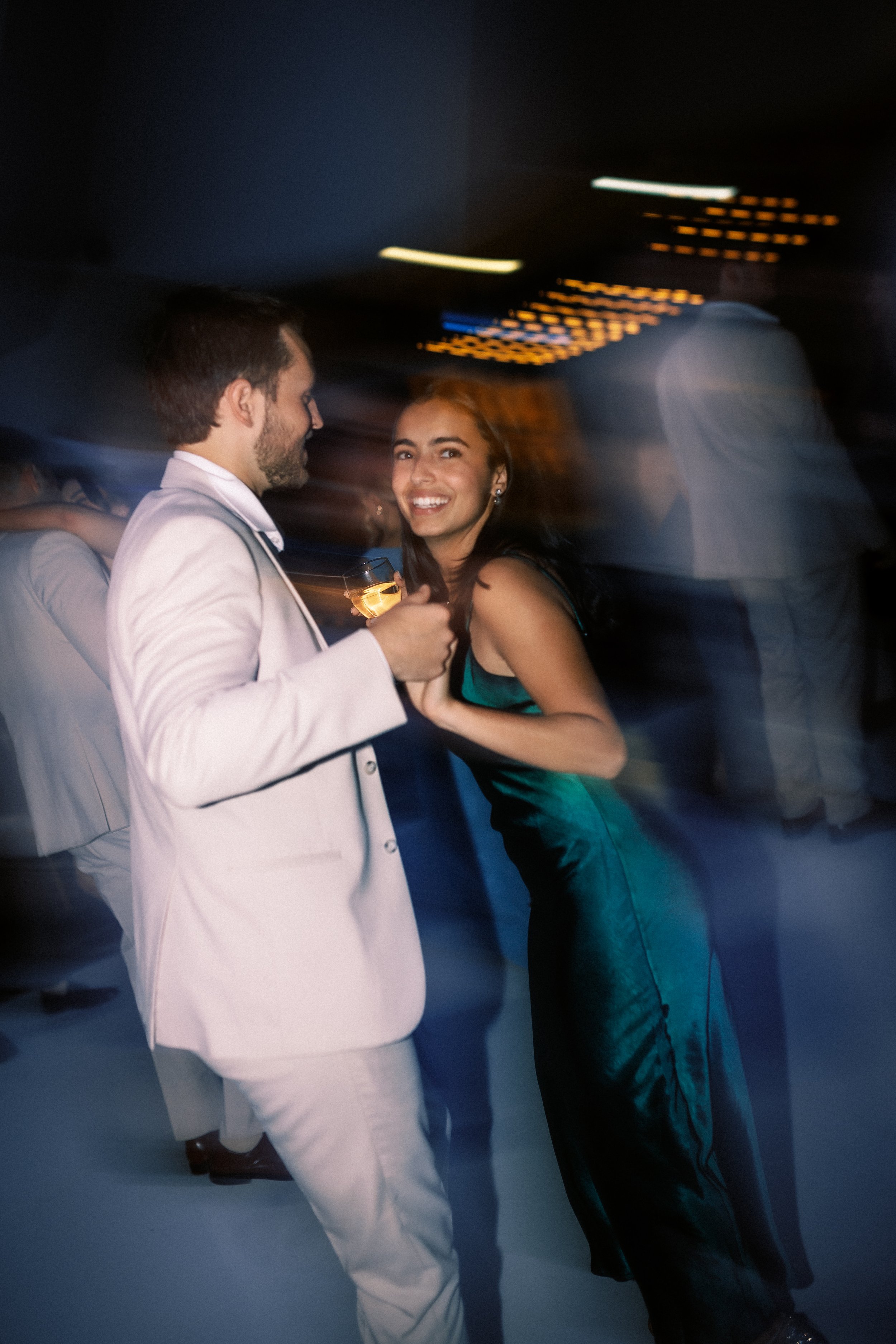 A man and woman dancing at a party or nightclub, with the man wearing a light-colored suit and the woman in a dark satin dress, both smiling and holding drinks, surrounded by blurred lights and other people.
