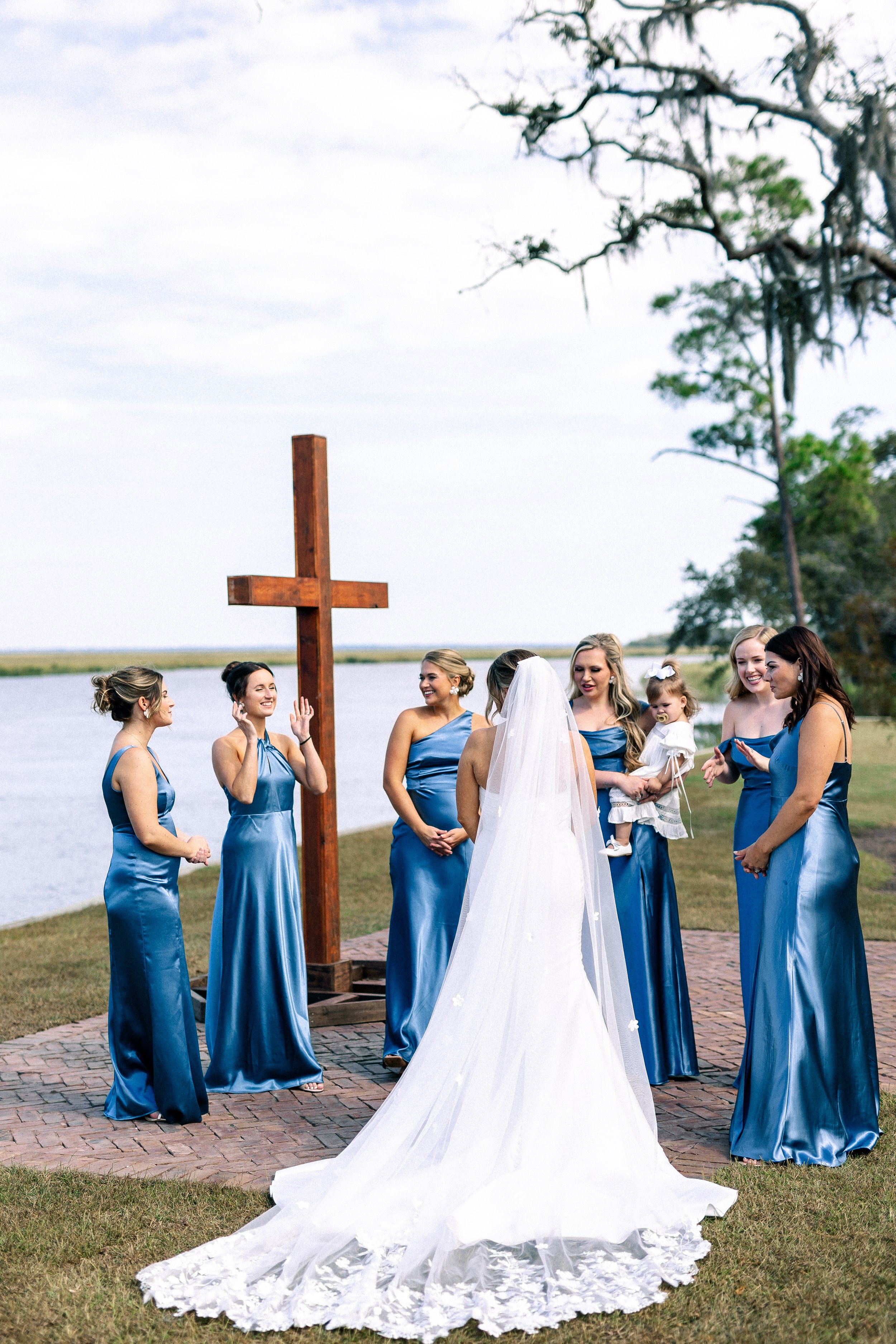 A bride and her wedding party standing outdoors near a large wooden cross, with water and trees in the background. The bridesmaids are wearing matching blue dresses, and the bride is in a white gown with a long veil, facing each other during a weddin