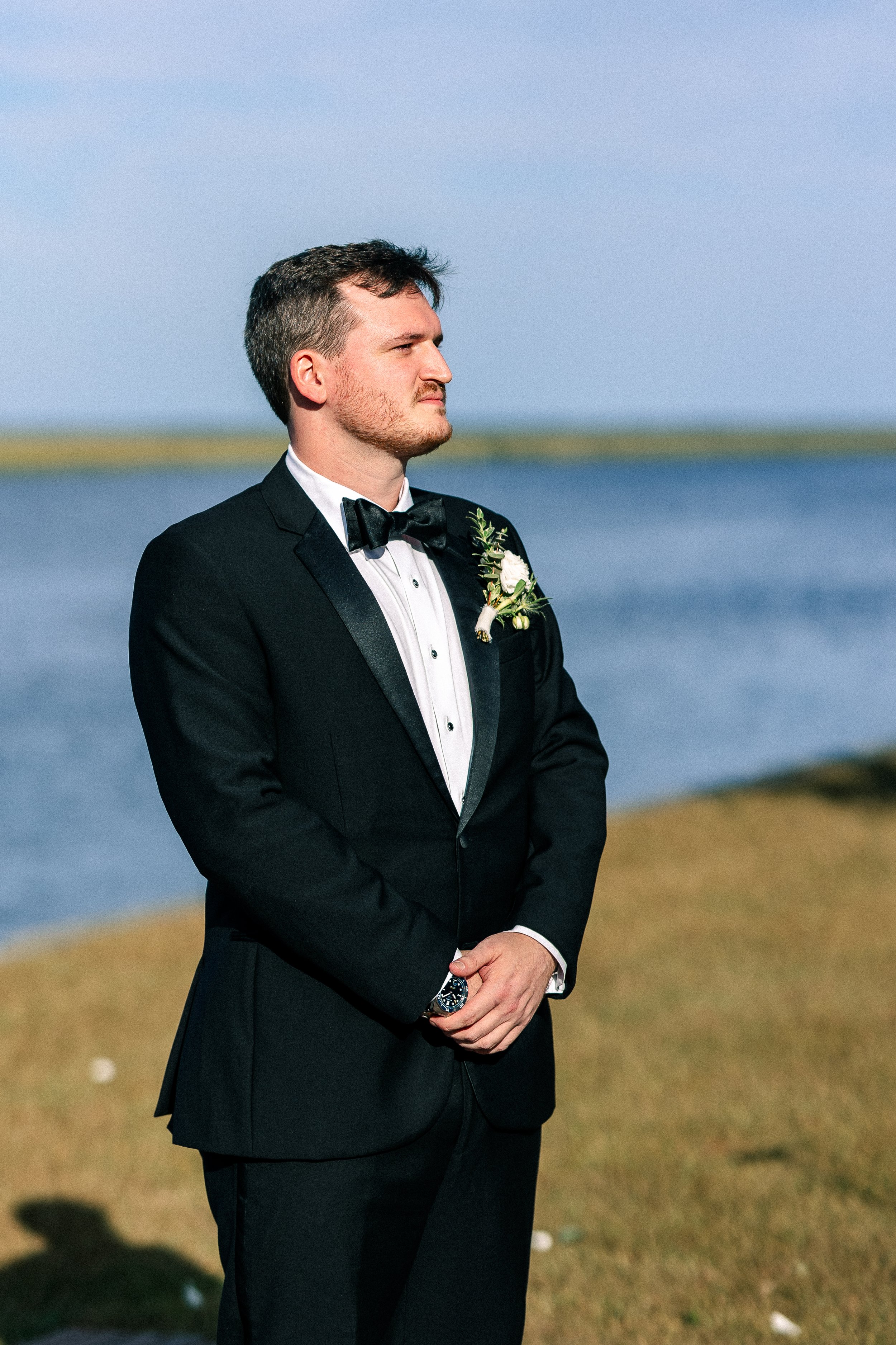 Man in a tuxedo with a boutonniere, standing outdoors near water, looking contemplative.