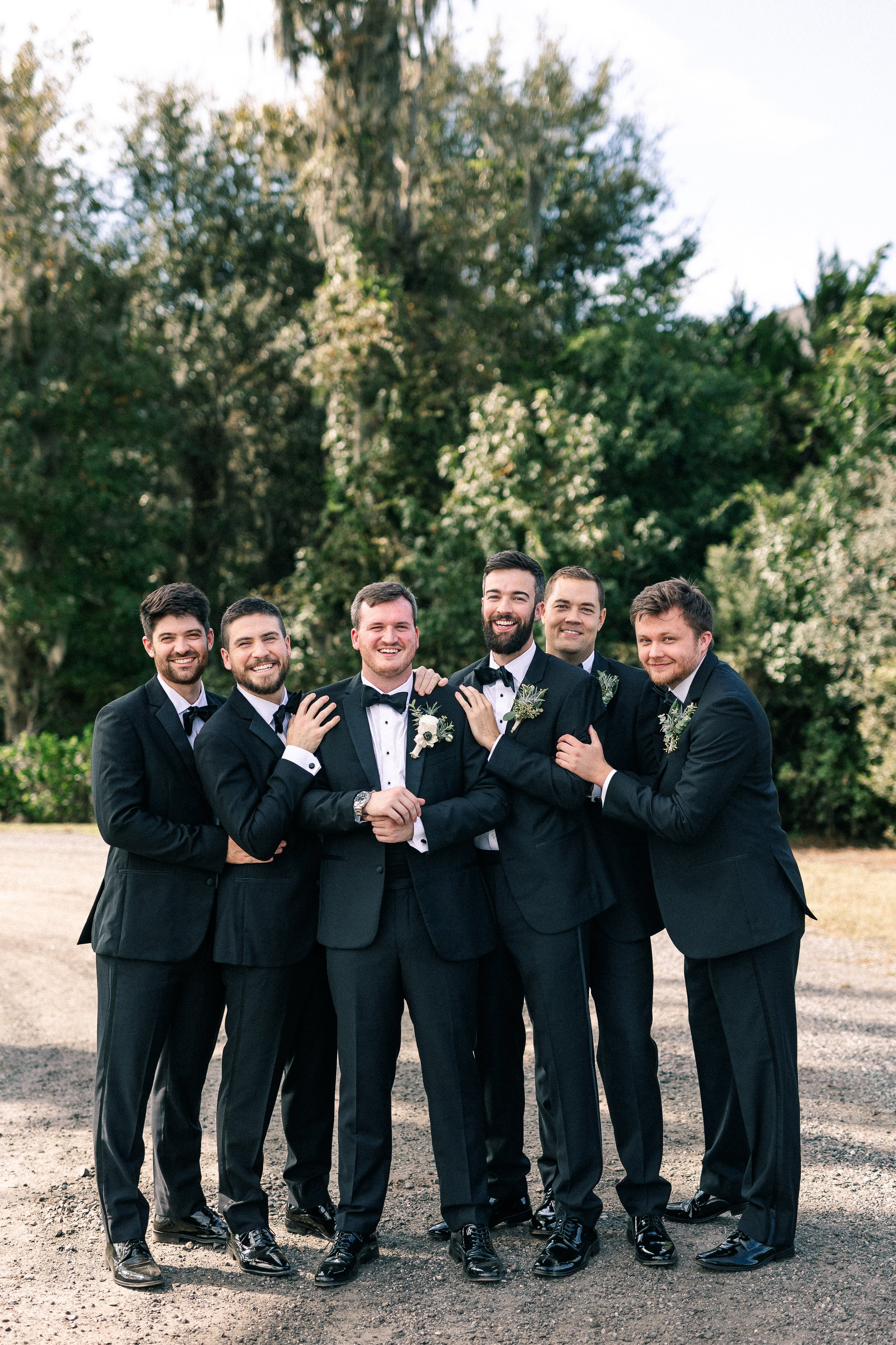 Group of six men dressed in black tuxedos with bow ties, standing outdoors on a gravel surface, smiling and embracing each other, with green trees and clear sky in the background.
