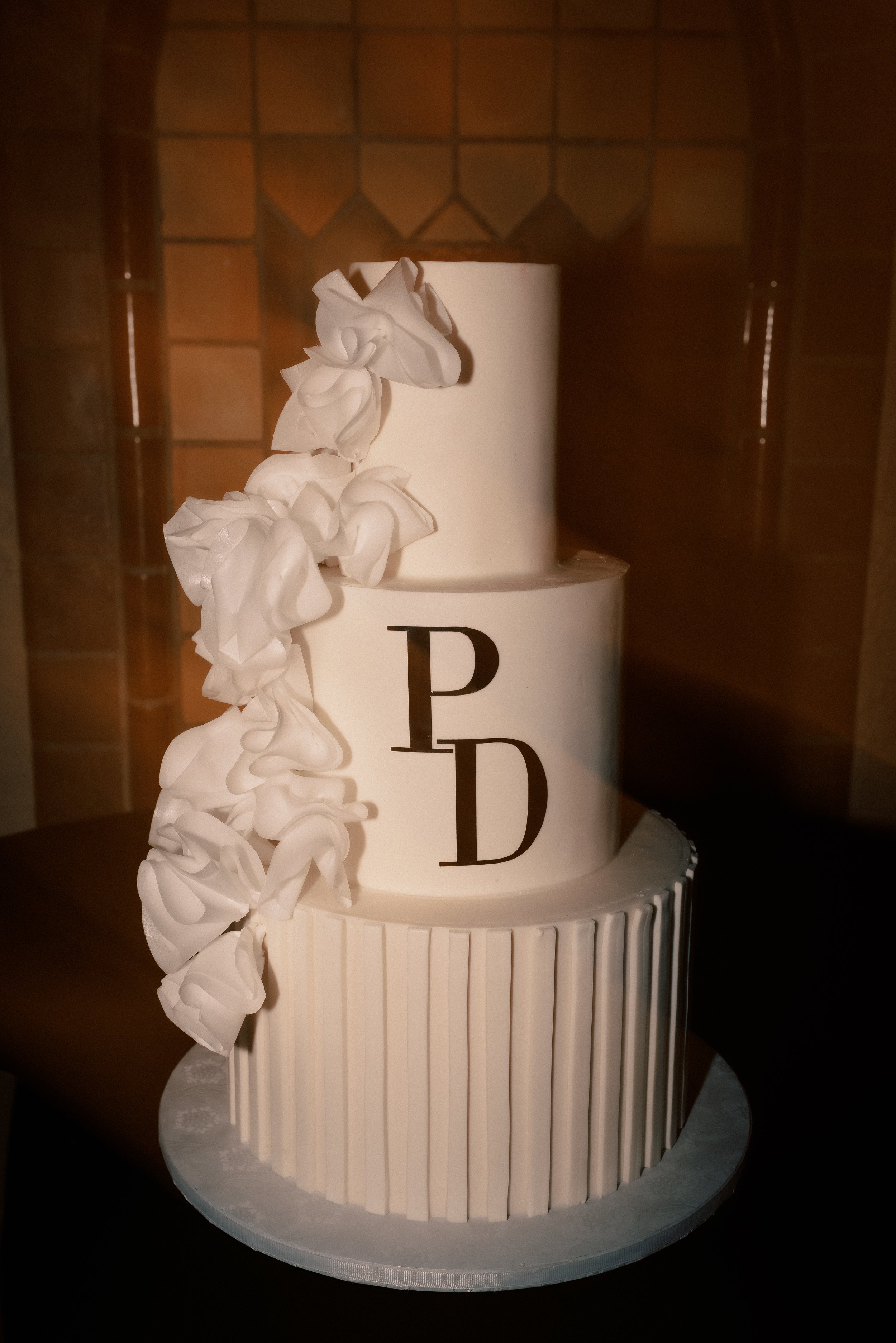 A three-tier white wedding cake with vertical striped design on the bottom tier, plain middle and top tiers, decorated with white paper flowers cascading down the side, and decorated with black letters 'P' and 'D' on the middle tier, placed on a roun