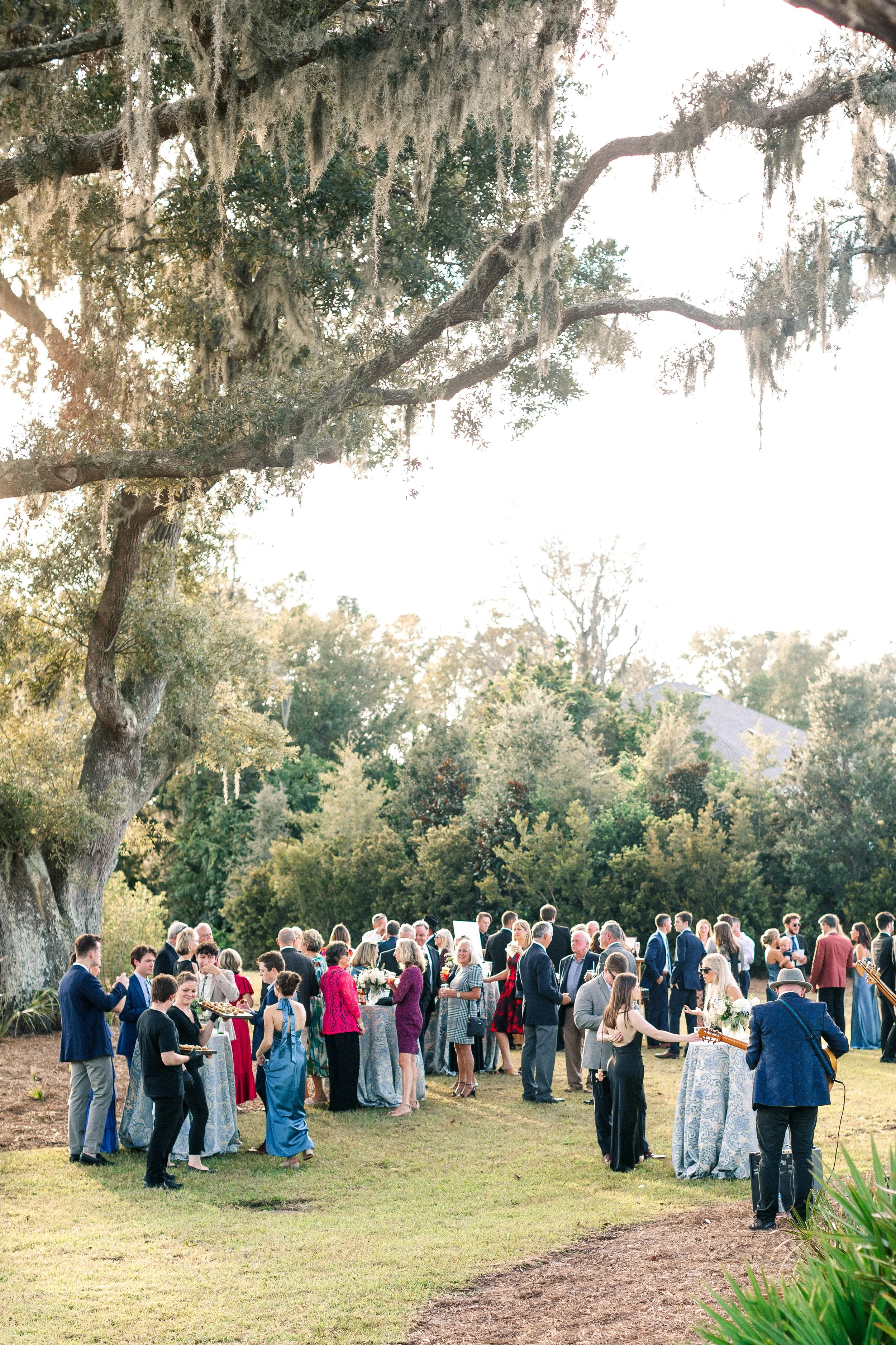 Outdoor gathering of people at a celebration or event, with some holding drinks, under large trees on a sunny day.