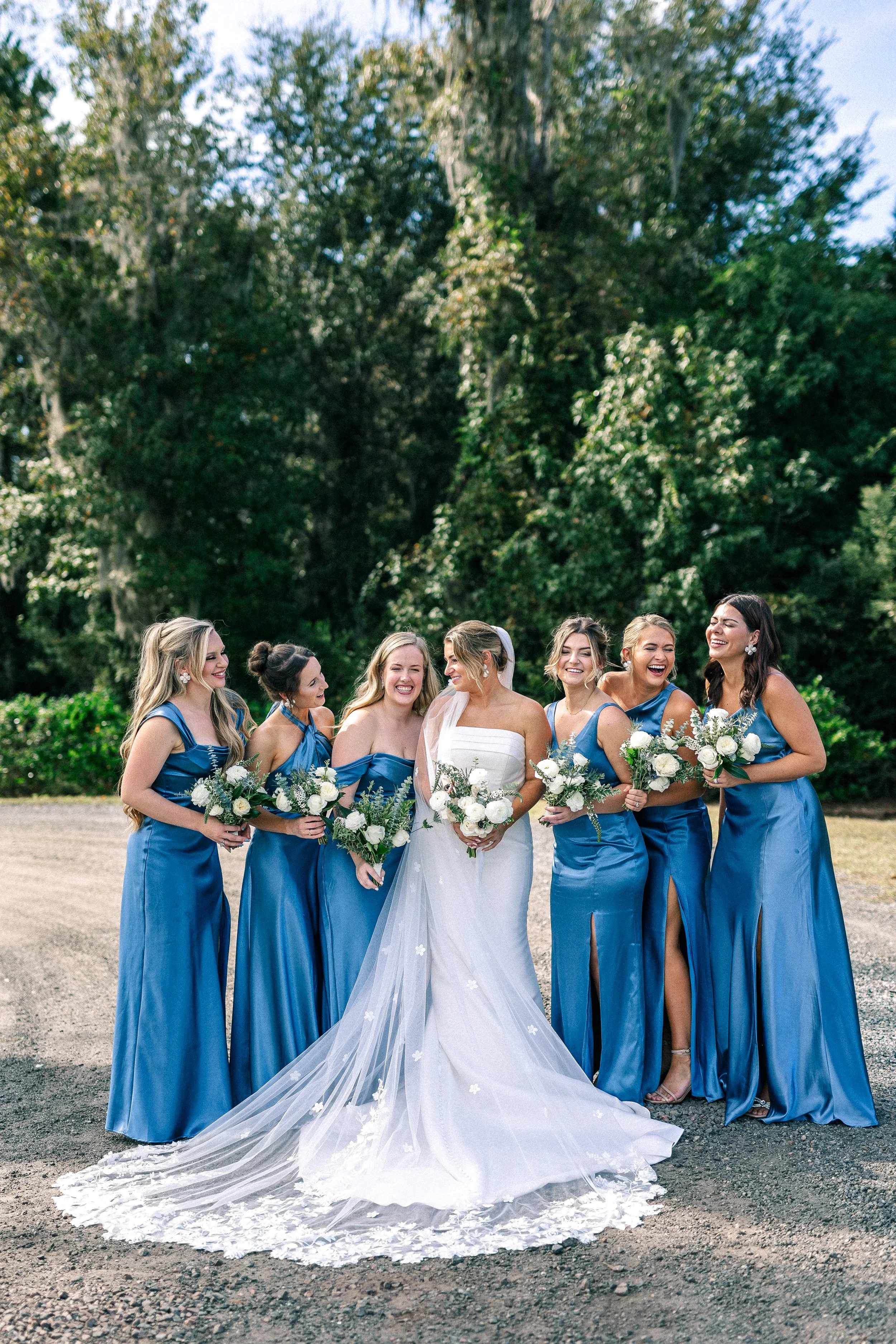 A bride in a white wedding gown holding a bouquet of white flowers, standing with six bridesmaids dressed in matching blue satin dresses, outside on a gravel area with green trees in the background.