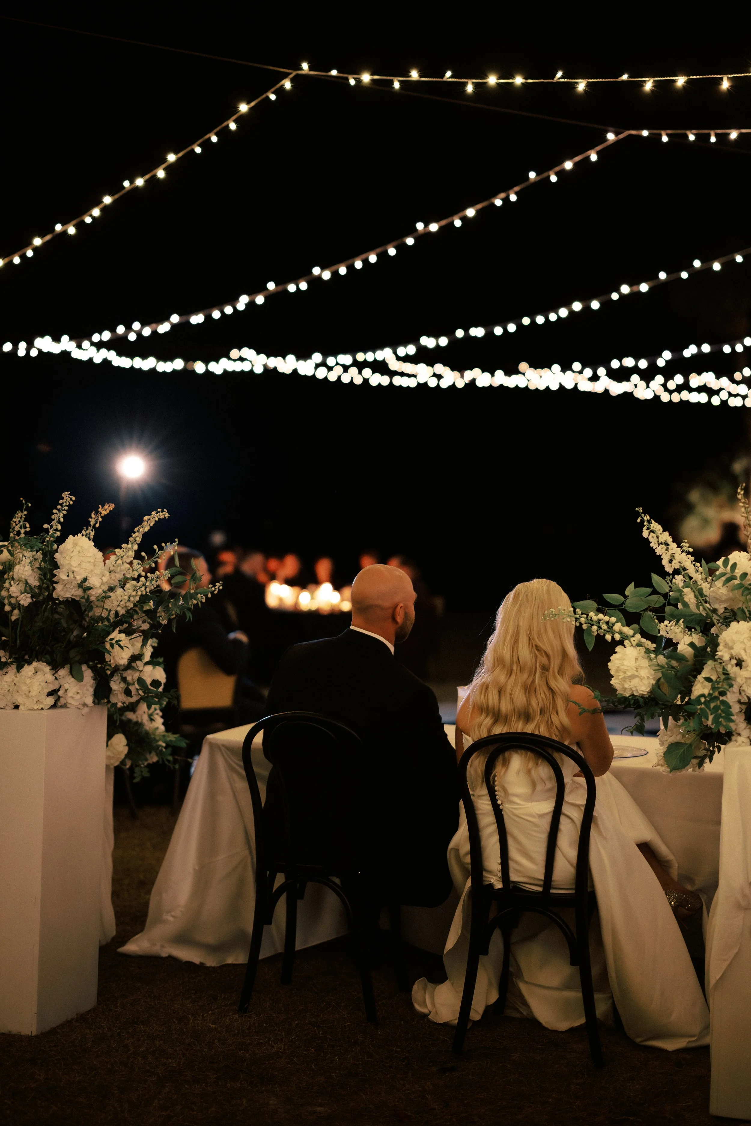 A couple dressed in formal attire sitting at a decorated table outdoors at night, illuminated by string lights overhead, with floral arrangements on the table and blurred guests in the background.