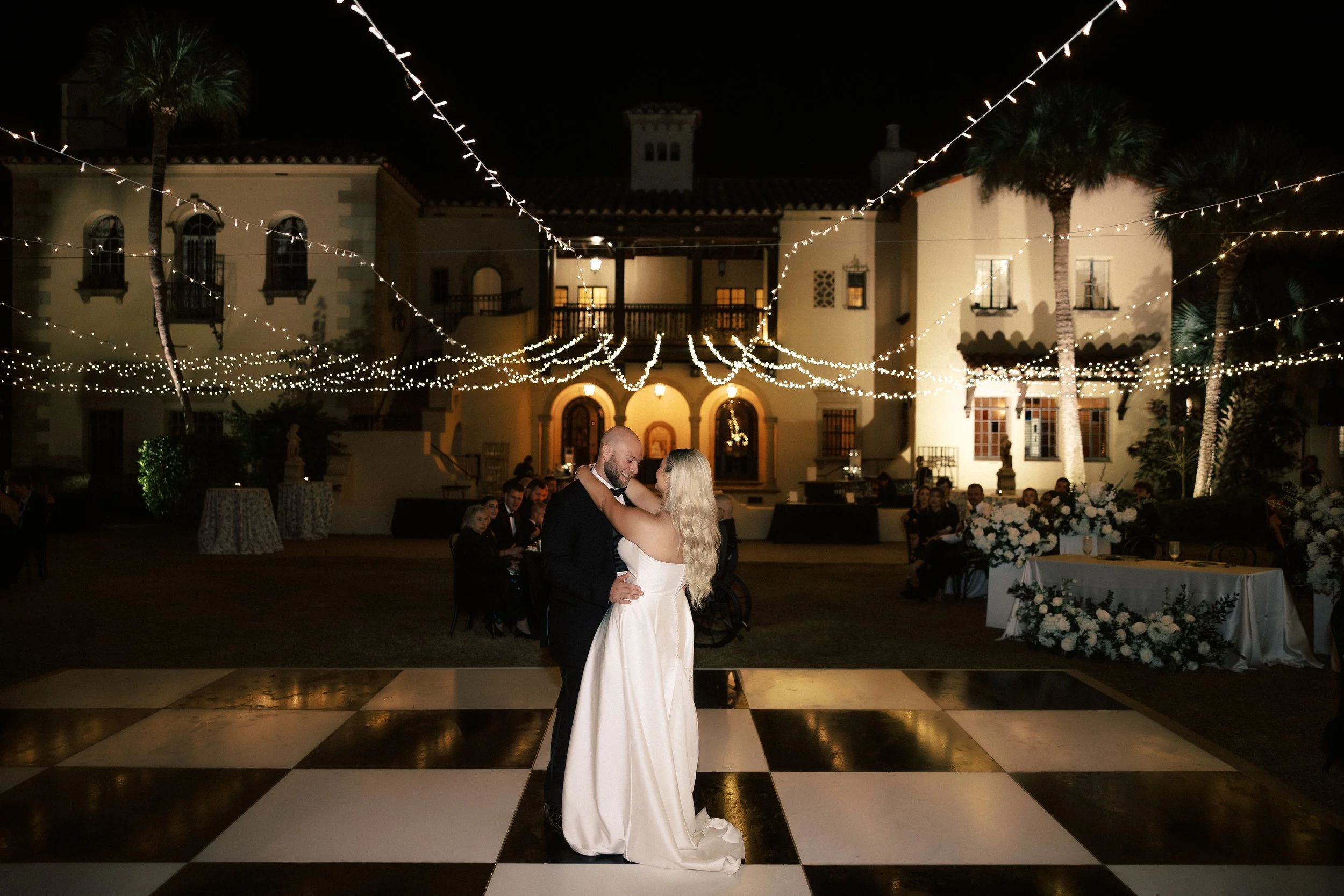 Bride and groom dancing on a checkered dance floor at night, illuminated by string lights, with wedding guests and florals in the background outside a grand house.