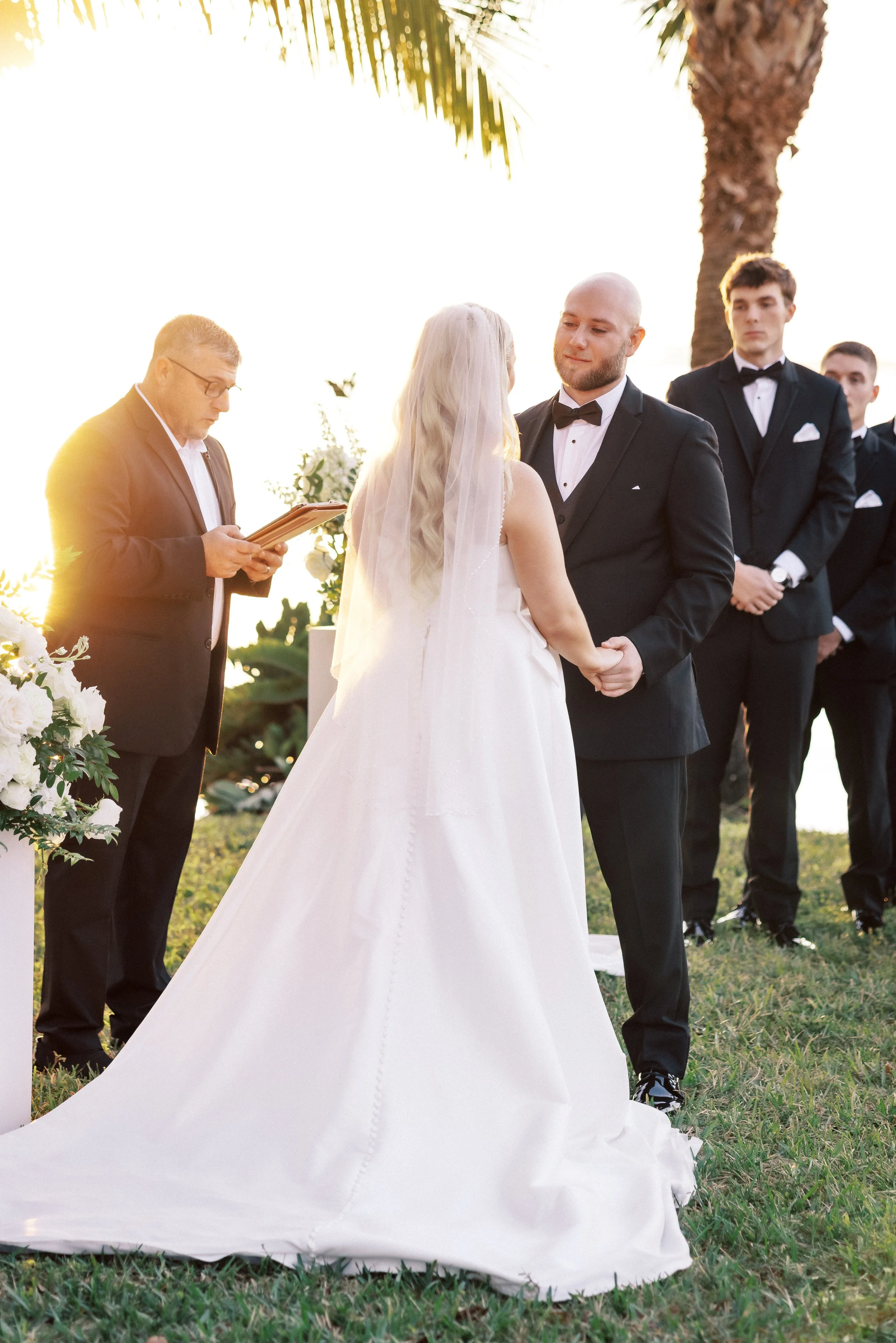 Bride and groom holding hands during outdoor wedding ceremony at sunset, officiant reading from a book, surrounded by groomsmen in suits, palm trees in background.