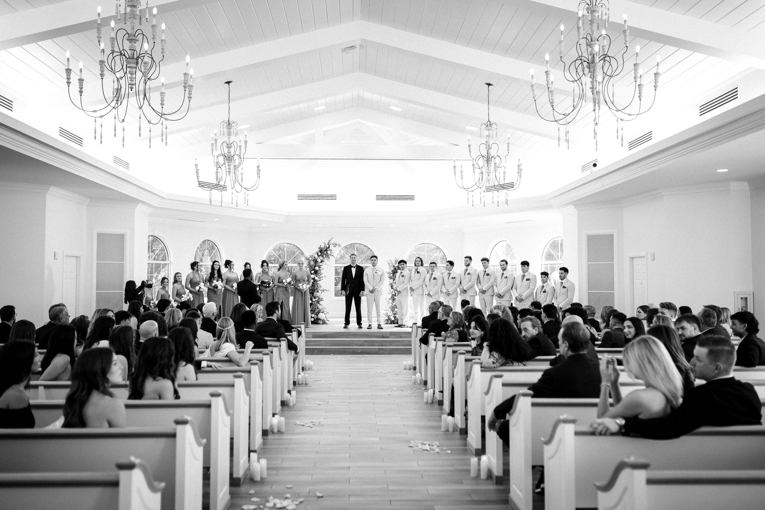 Black and white photo of a wedding ceremony in a bright, spacious hall with chandeliers, a stage with the bride and groom, bridal party standing on either side, and guests seated in pews watching.
