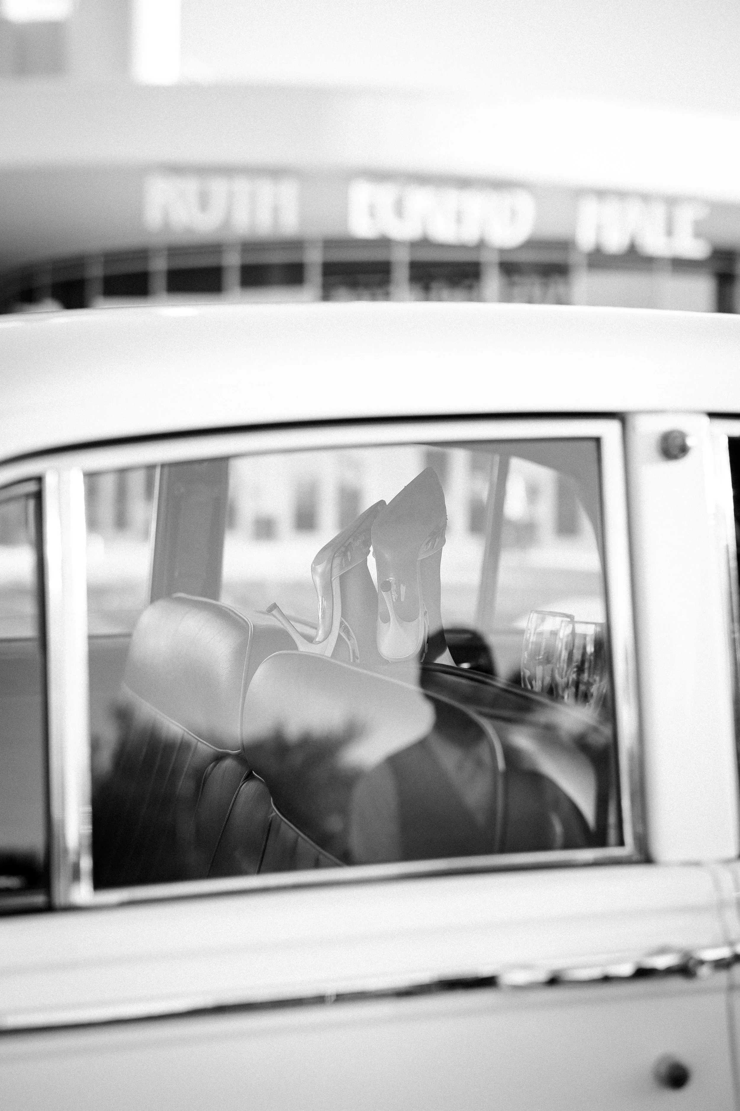 Black and white photo of the interior of a vintage car with a pair of high-heeled shoes on the front seat, seen through the window. The background includes a building with a sign that reads 'NORTH COND PLE'.