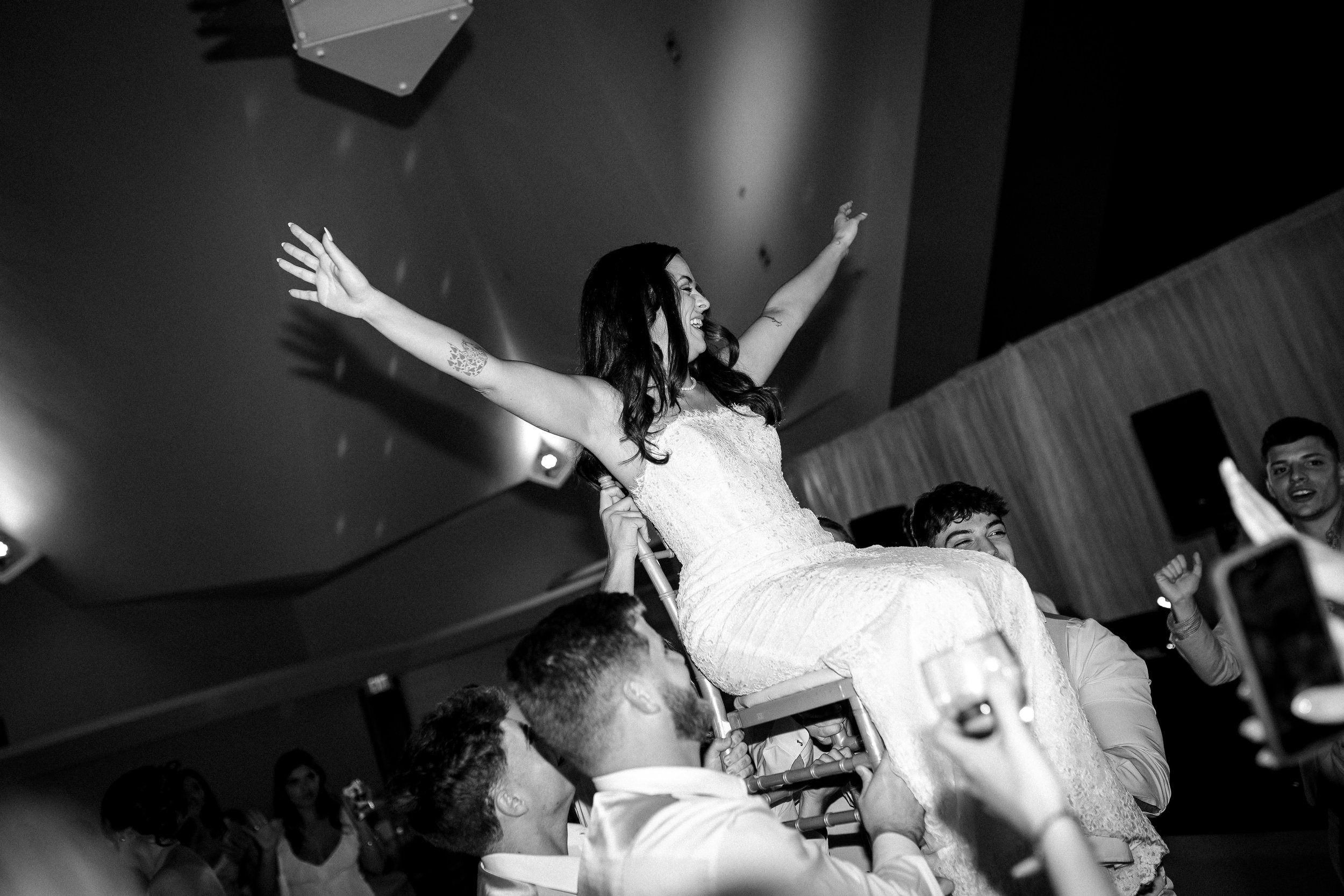Bride in a wedding dress being lifted in a chair during her celebration, arms outstretched, laughing and surrounded by guests.