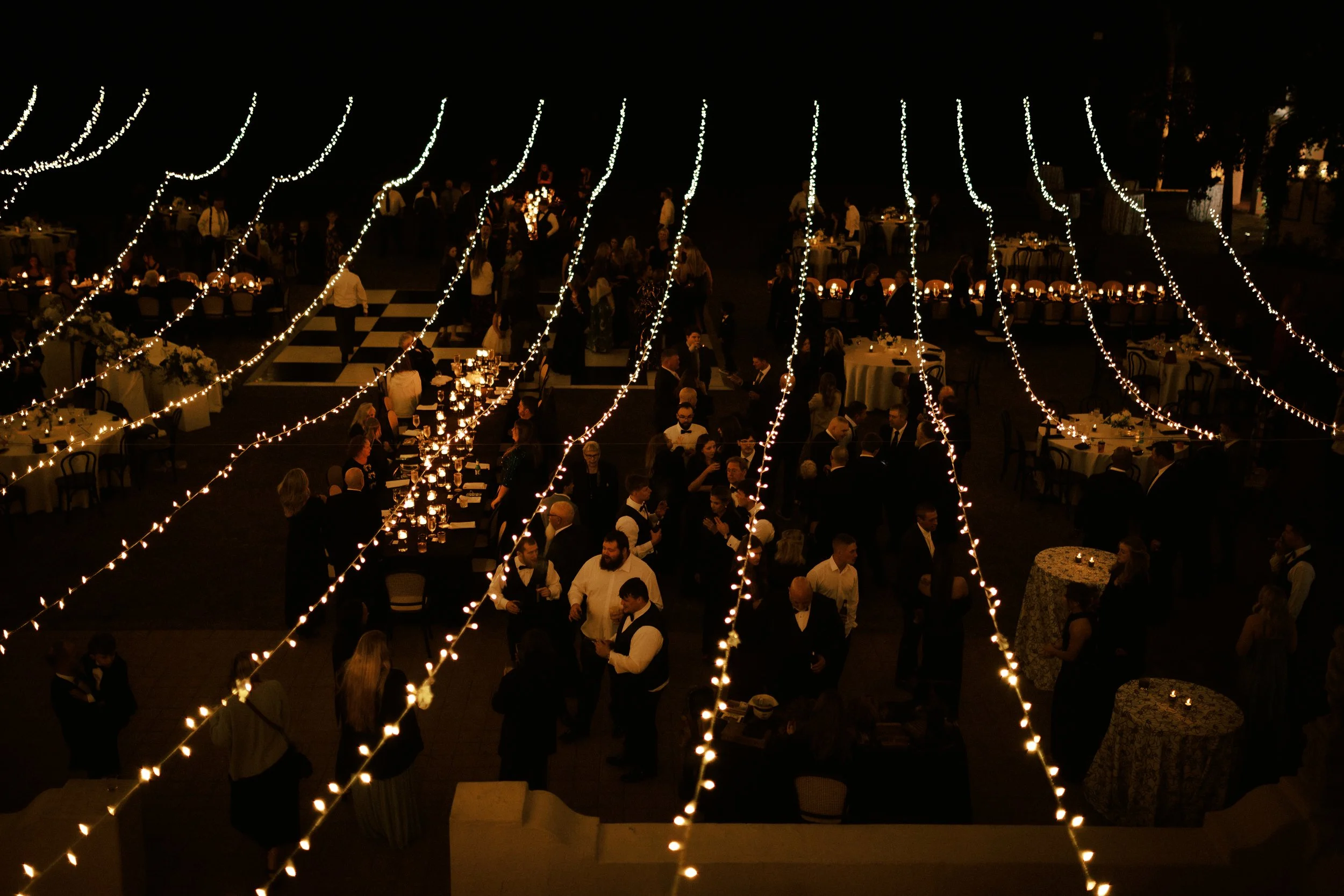 Nighttime outdoor party with string lights, people socializing, and decorated tables.