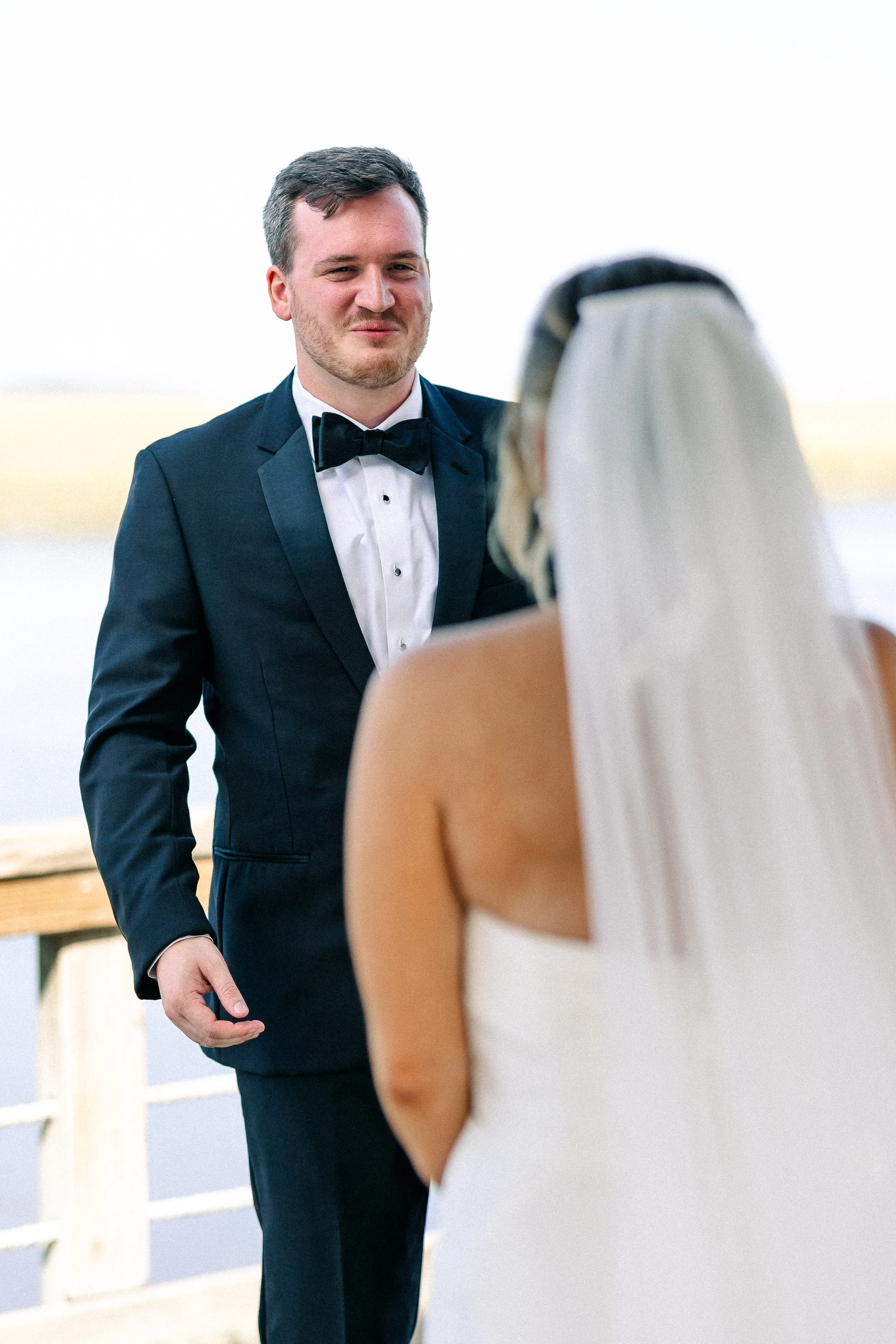 A groom in a black tuxedo and bow tie weds a bride in a white wedding dress on a dock near the water.