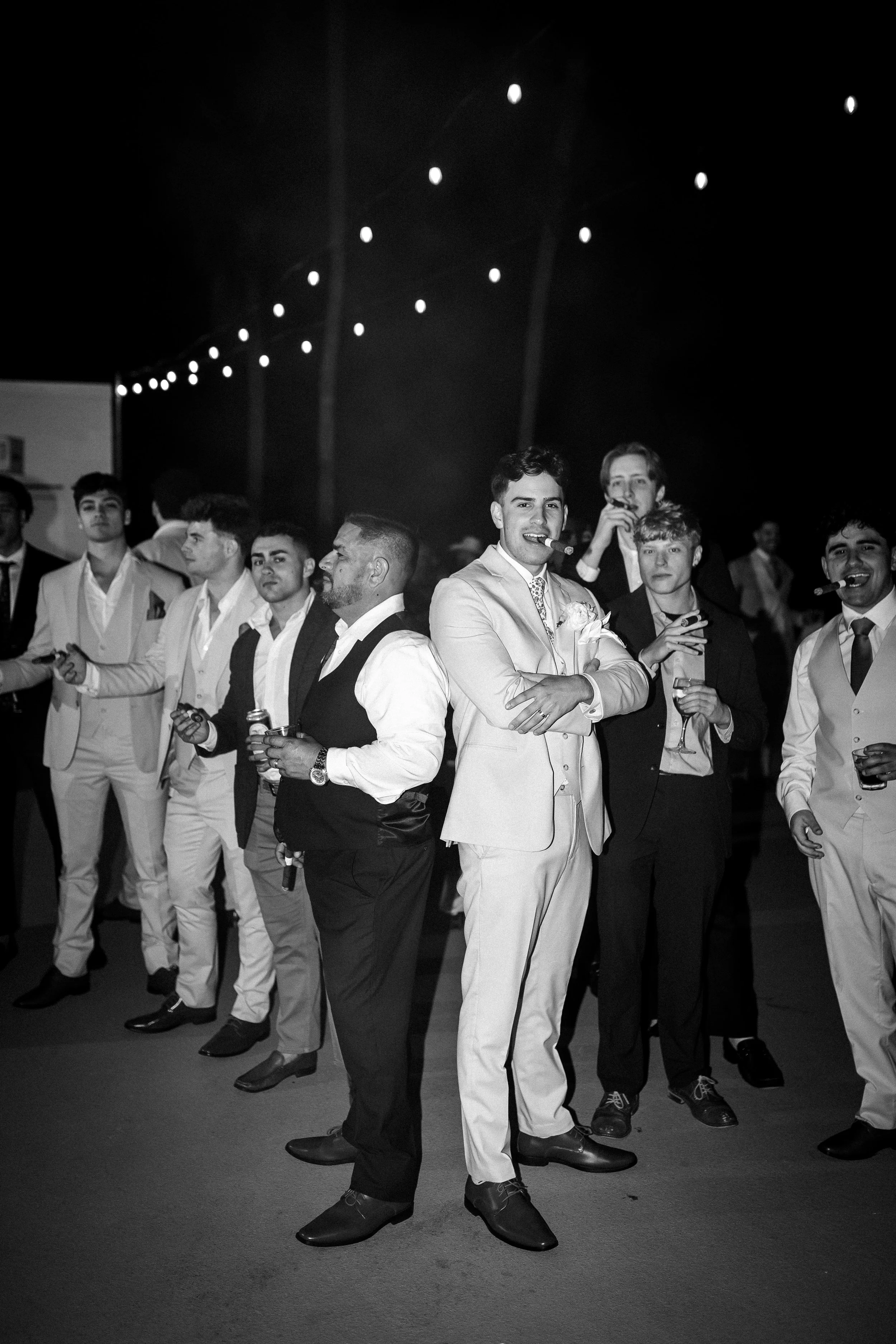 Group of men dressed in suits at night, some smoking cigars, under string lights, at a celebration or party.