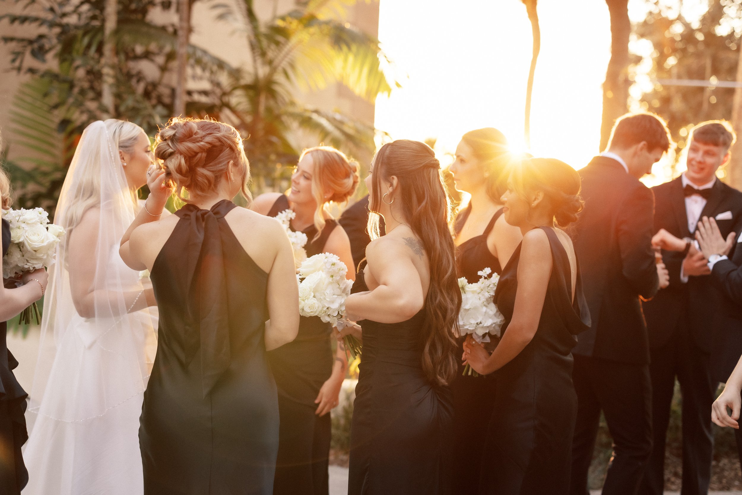 A group of women in black dresses and men in tuxedos at an outdoor wedding reception during sunset, exchanging flowers and conversation.