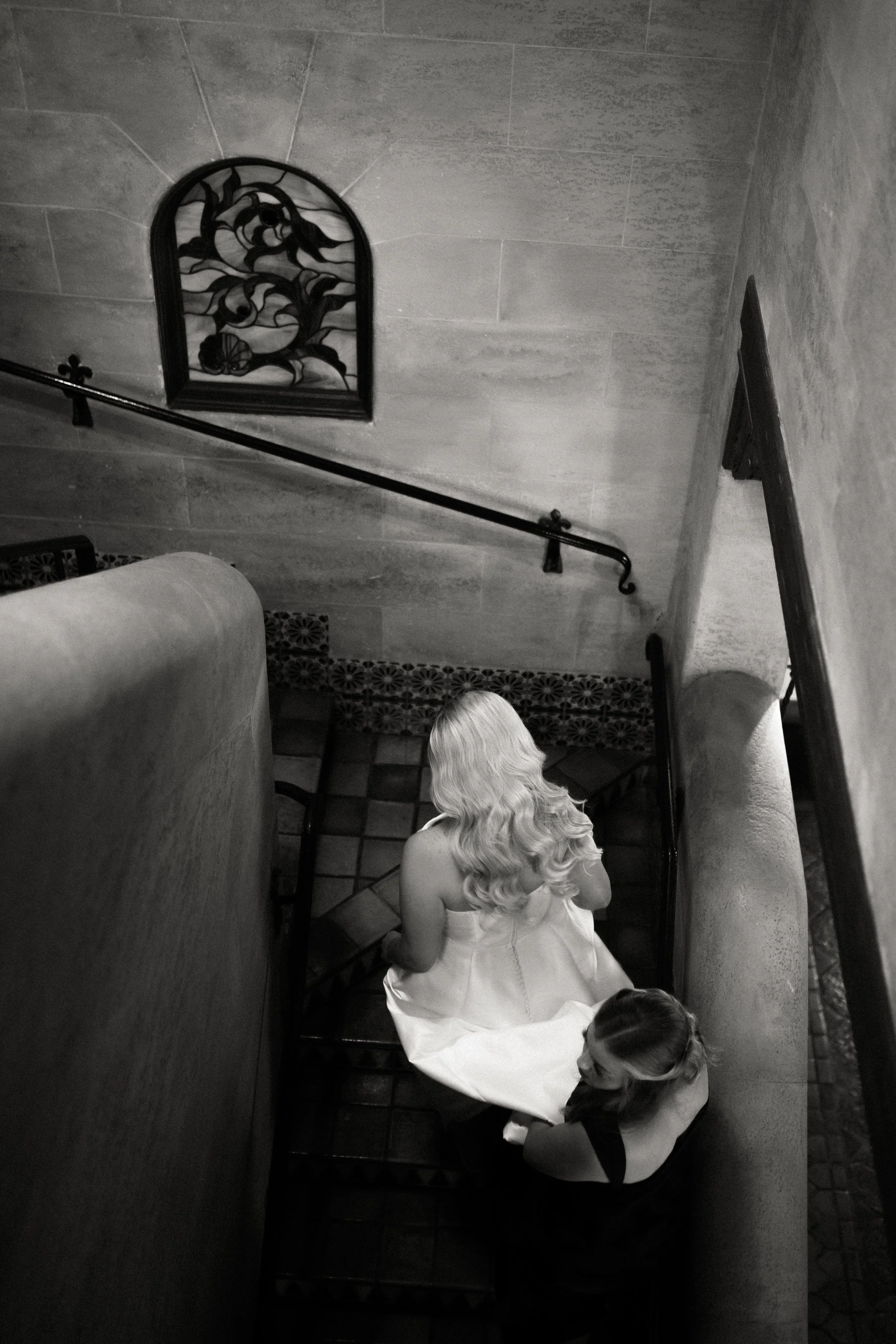 A woman in a white dress with long blonde hair sitting on stairs, viewed from above, with another woman in dark clothing standing nearby, in a setting with tiled stairs, a wrought iron decorative wall piece, and stone walls.