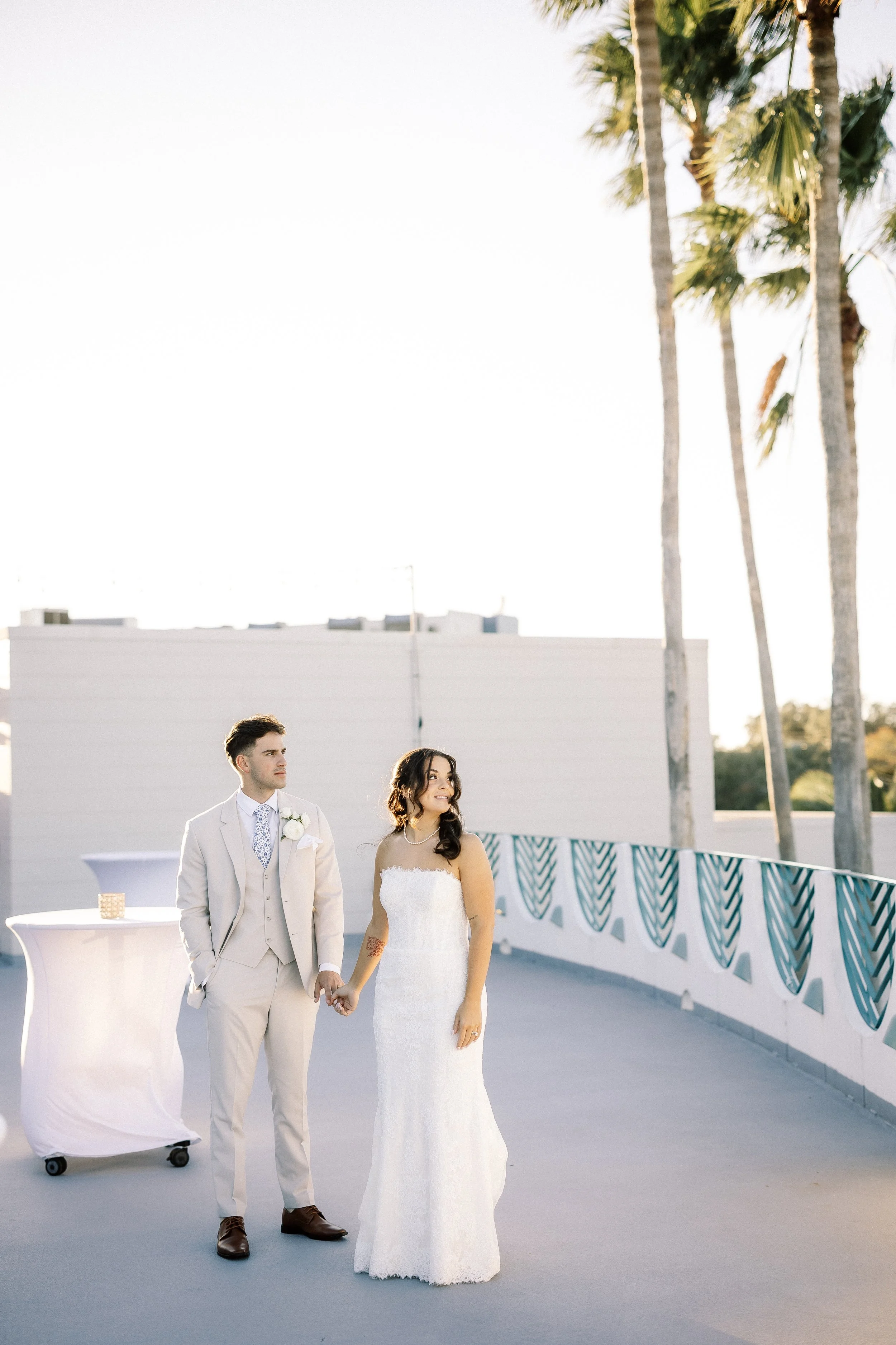 A bride and groom holding hands on a rooftop, with palm trees and a bright sky in the background, during a wedding ceremony.