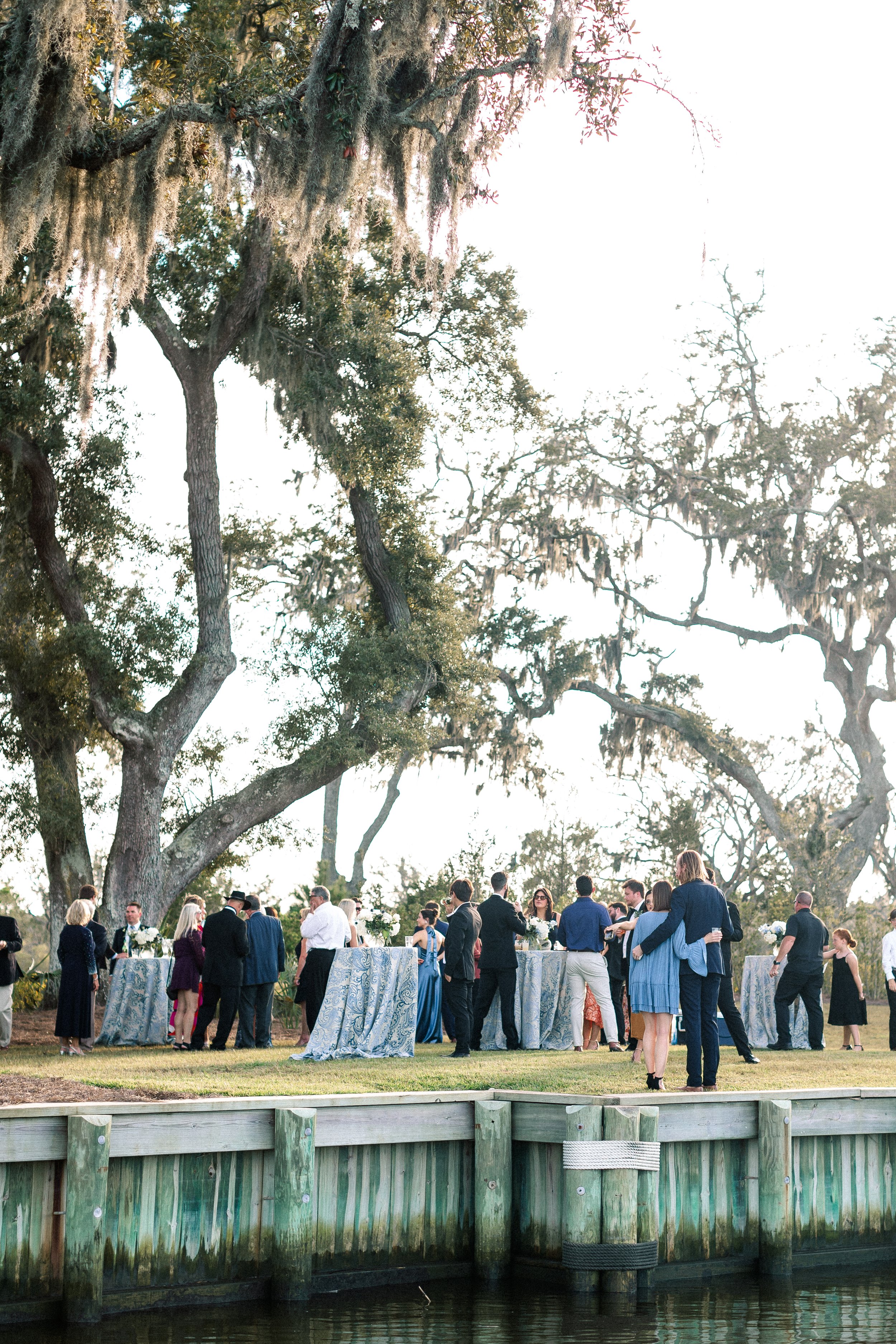 A group of people dressed in formal attire are gathered outdoors at a social event, standing around several tall tables with blue patterned tablecloths, set among large trees with Spanish moss, near a body of water with a wooden dock.