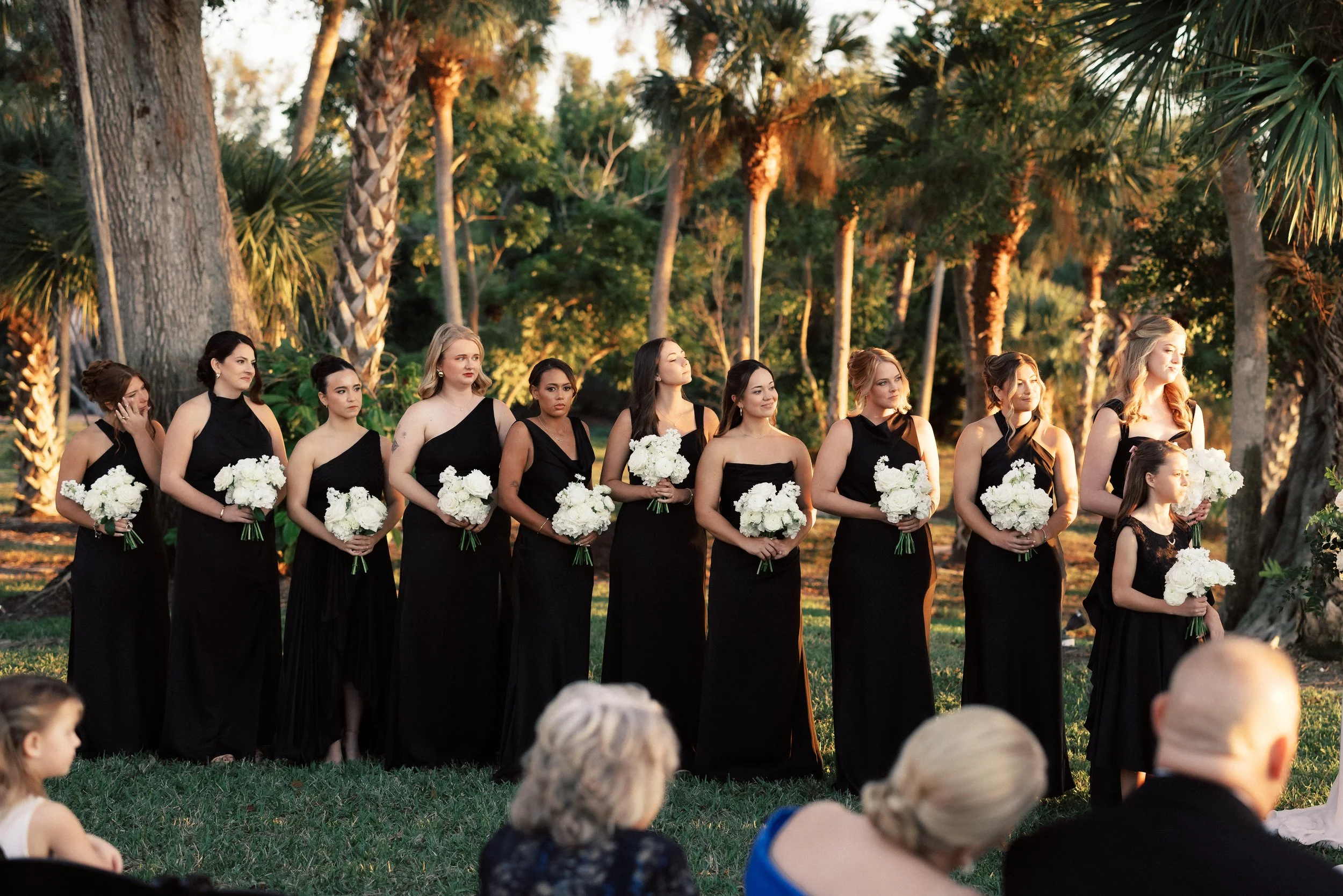 A group of women in black dresses holding white flower bouquets in an outdoor setting with trees, possibly during a wedding or formal event.